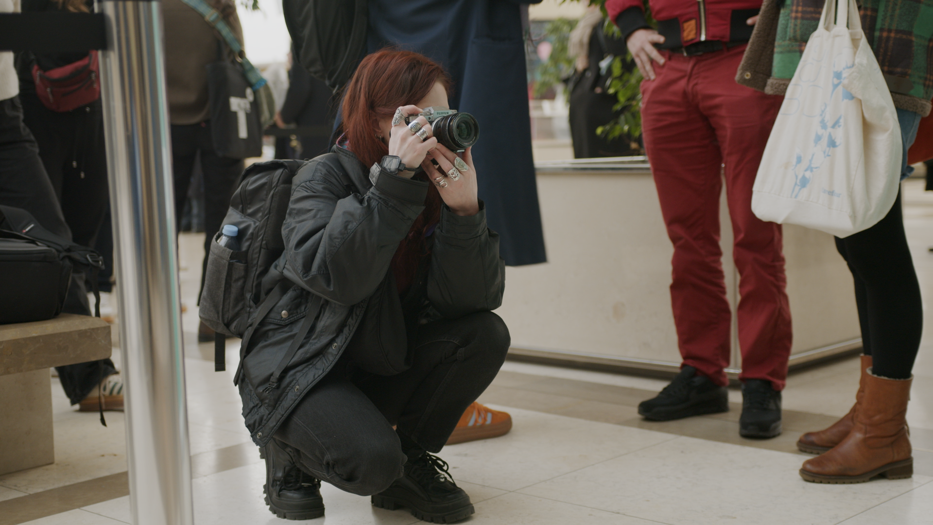 Une personne avec des cheveux rouges, portant une veste noire, regarde à travers l'objectif d'un appareil photo numérique, en position accroupie, dans un lieu public où plusieurs personnes se tiennent debout.