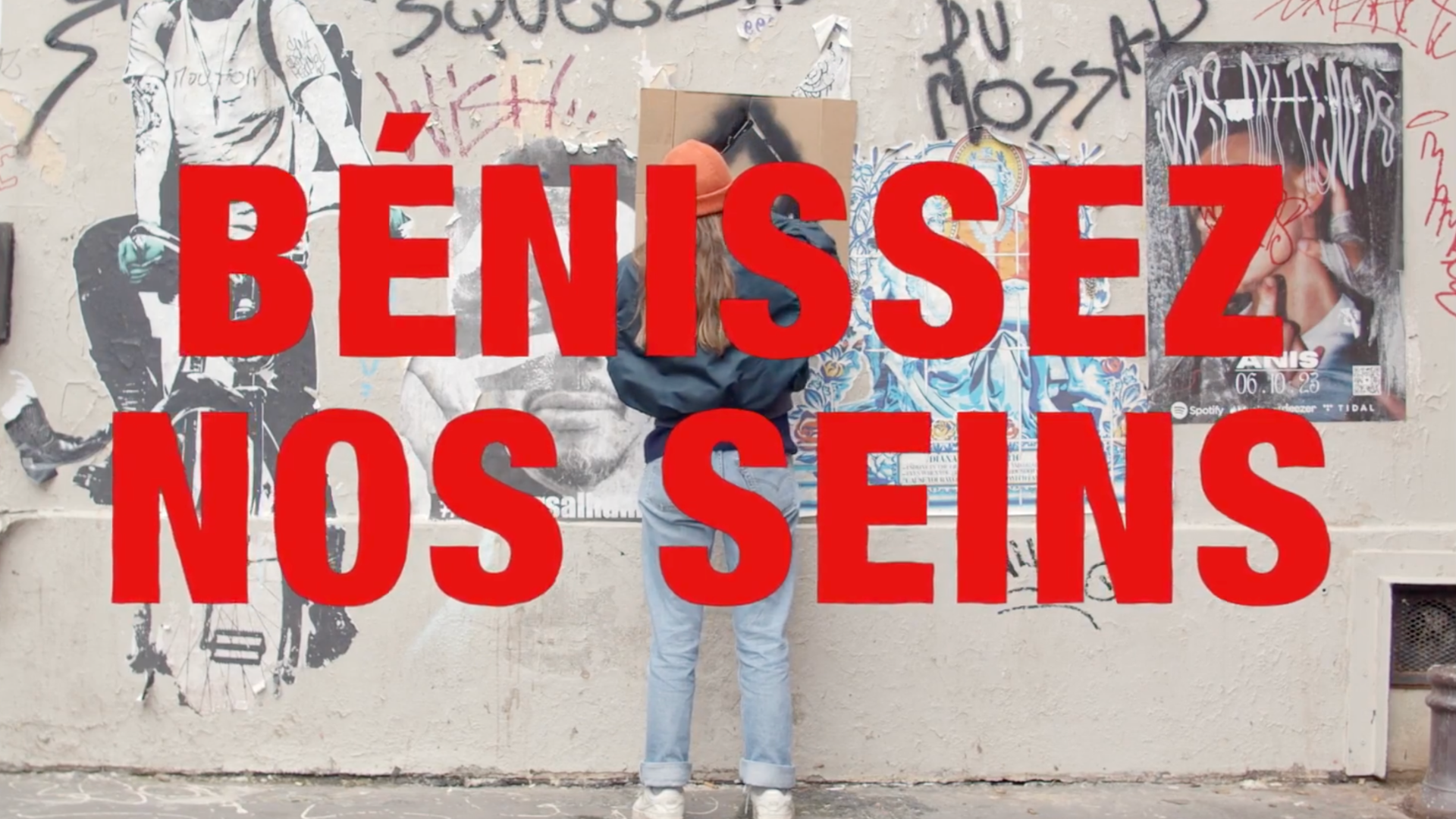 Une personne avec des cheveux longs, portant une veste noire, un chapeau orange et un jean, elle se tient face à un mur couvert de graffitis et d'affiches. La photo est superposée d'un message en gros caractères rouges disant "BÉNISSEZ NOS SEMS".