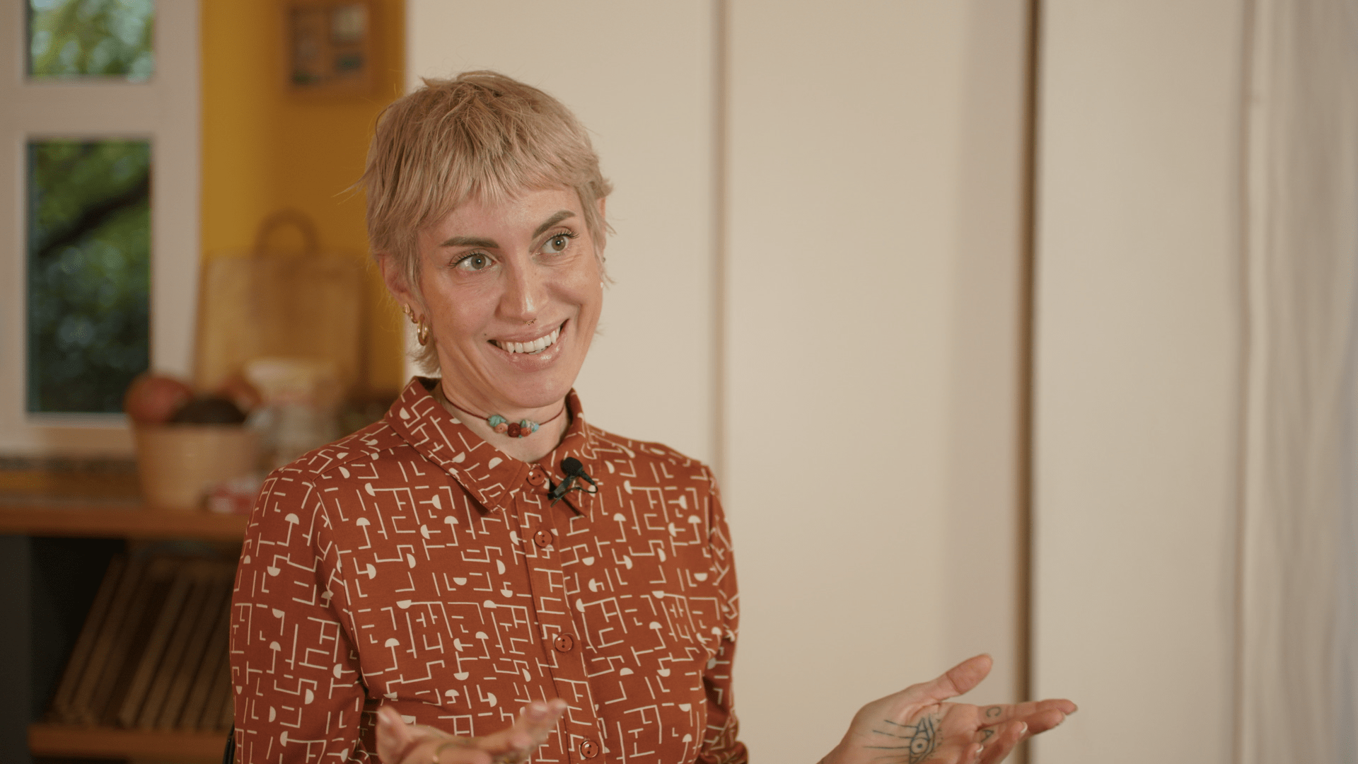 Une femme aux cheveux courts et blonds, portant une chemise rouge avec un motif blanc, parle avec un sourire dans une pièce chaleureuse et décorée, tenant peut-être un discours ou étant interviewée.