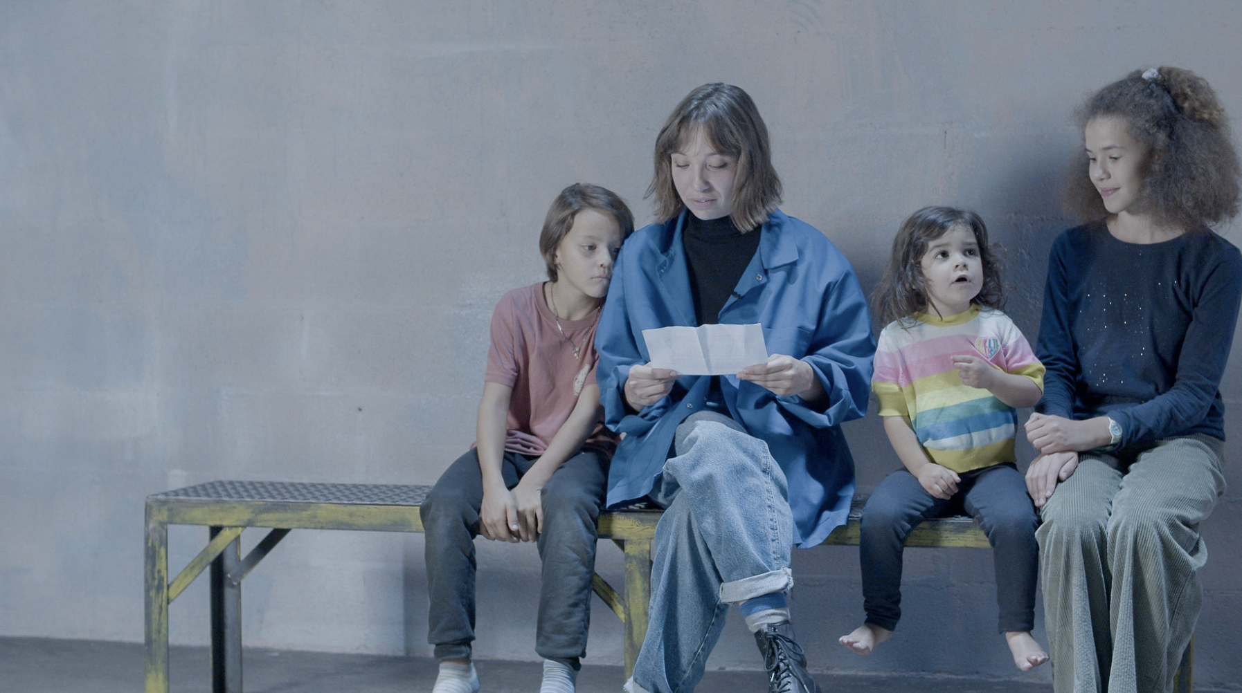Quatre personnes assises sur un banc en metal contre un mur gris, une femme avec une lettre à la main et trois enfants autour, l'un d'eux pointant quelque chose et une autre regardant la femme.