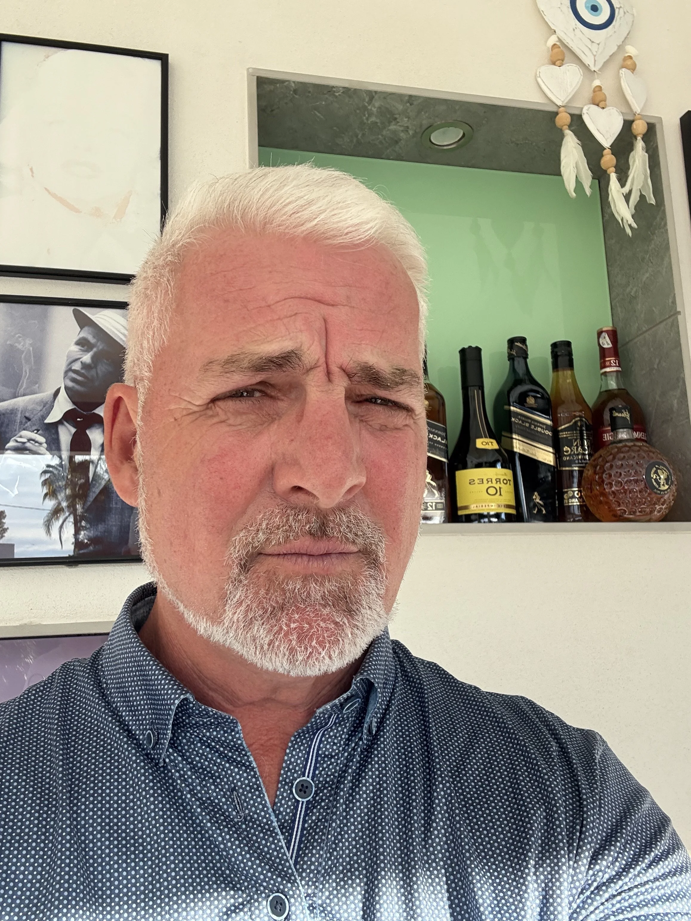 A man with white hair and a beard takes a selfie indoors. Behind him, on a shelf, are multiple bottles of alcohol including whiskey and liqueur. To the left, there is a black-and-white framed photograph or poster of a man in a suit and fedora hat. To the right, there is a wall decoration with white hearts, beads, and feather-like elements. The man is wearing a dark blue, polka-dotted shirt.