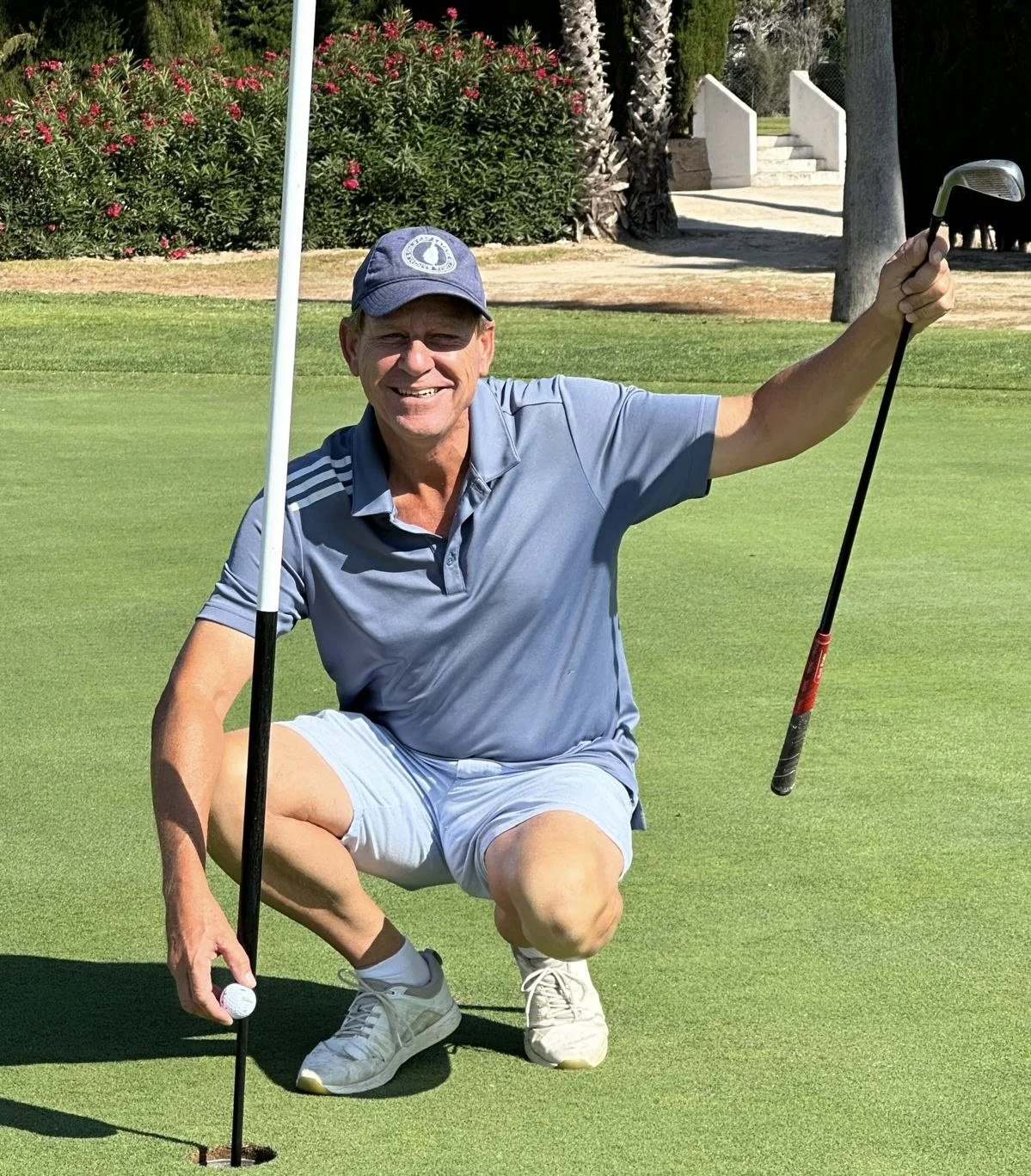 A smiling man in a blue polo shirt, light shorts, and a cap, crouching on a golf green, holding a golf club in one hand and a golf ball in the other, next to the hole.