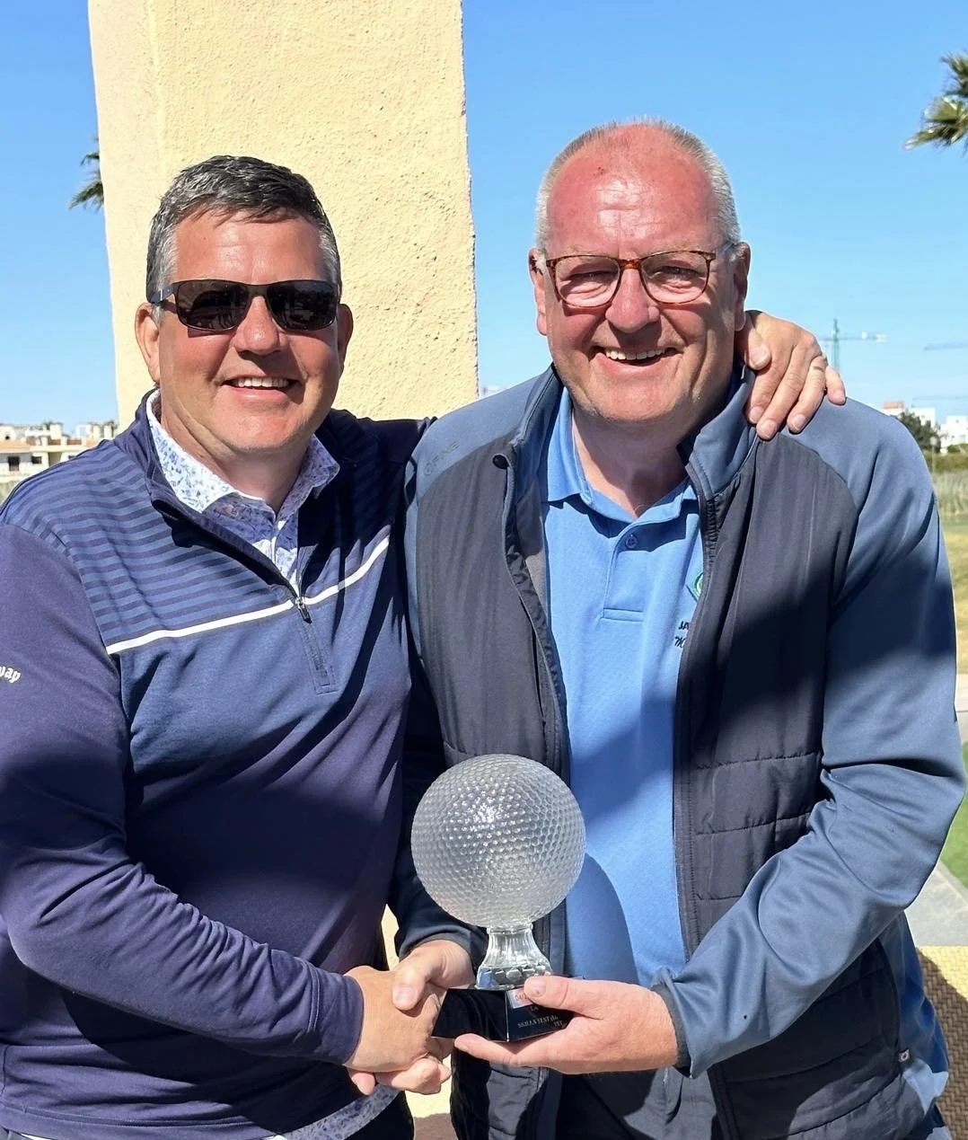 Two smiling men in golf attire holding a golf trophy together outdoors on a sunny day.