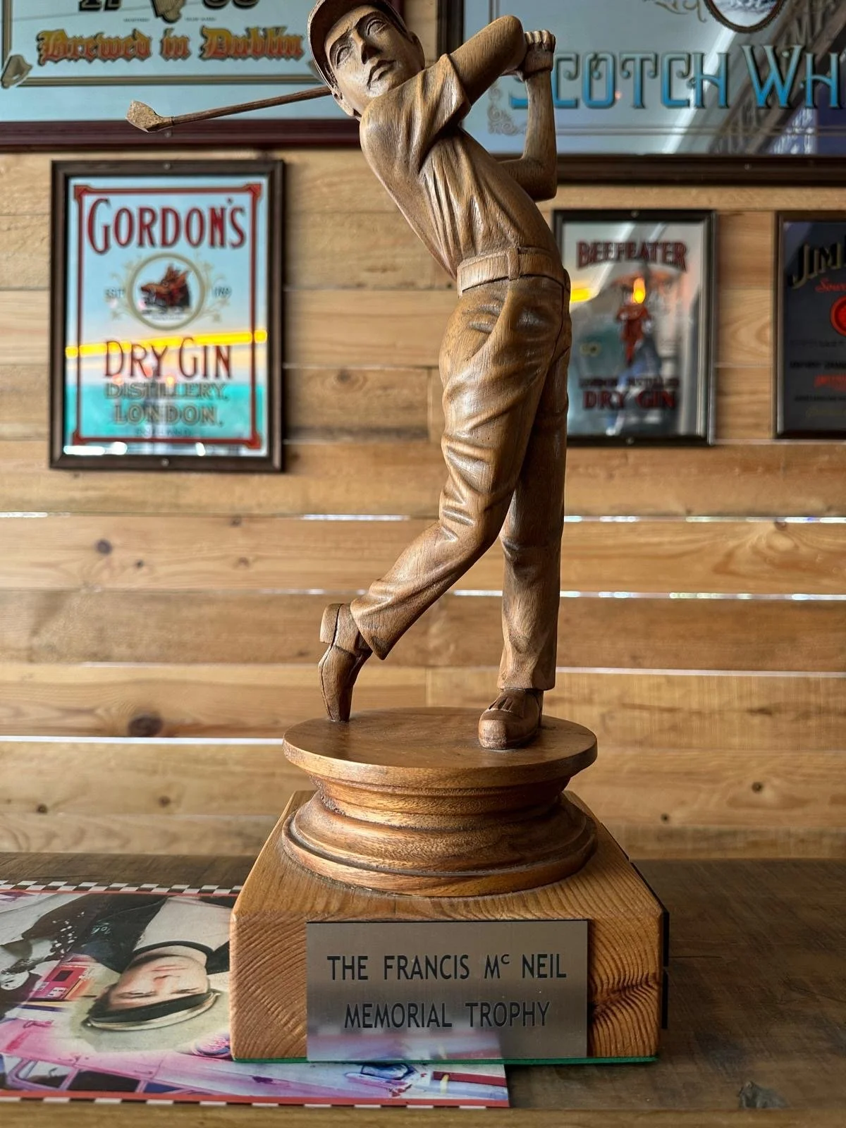 Wooden golf trophy of a golfer in mid-swing, with a plaque that reads 'The Francis Mc Neil Memorial Trophy', set on a wooden surface with a blurred background of vintage signs.