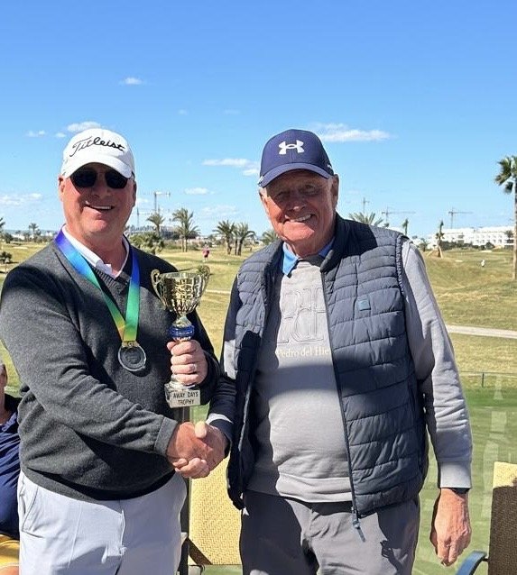 Two men smiling and shaking hands outdoors on a golf course, one wearing a white cap, sunglasses, and holding a trophy, the other wearing a navy cap and a gray vest, with a clear blue sky and palm trees in the background.