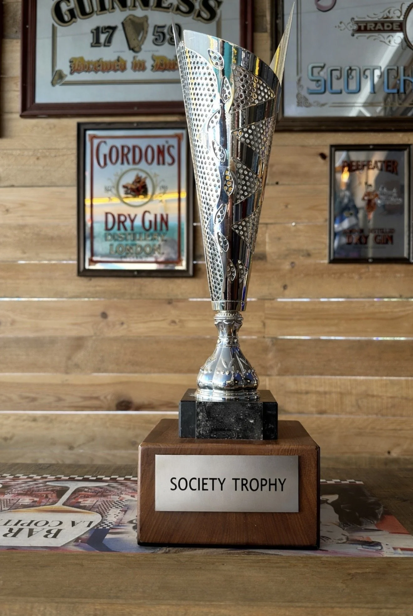 A silver trophy with a modern, intricate design on a black marble base, labeled 'Society Trophy,' placed on a wooden table in front of a wooden wall with framed advertisements for Guinness, Gordon's Dry Gin, and Scotch.