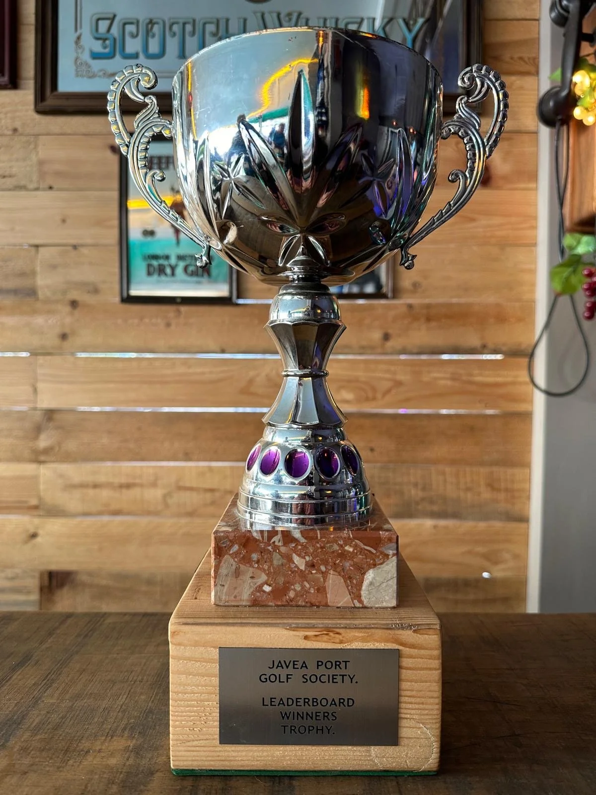 A silver trophy with ornate handles and decorative purple accents on its base, placed on a wooden stand with a metal plaque that reads: 'JAVEA PORT GOLF SOCIETY LEADERBOARD WINNERS TROPHY.' In the background, there is a wooden wall and a framed poster.