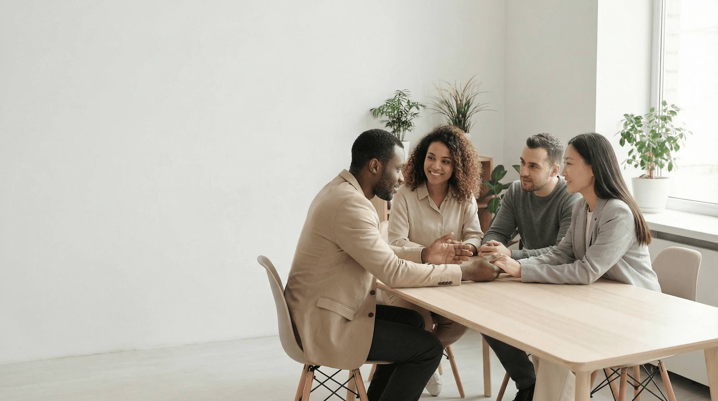 Four diverse people sitting around a wooden table, engaging in a warm conversation, with a bright room and potted plants in the background.