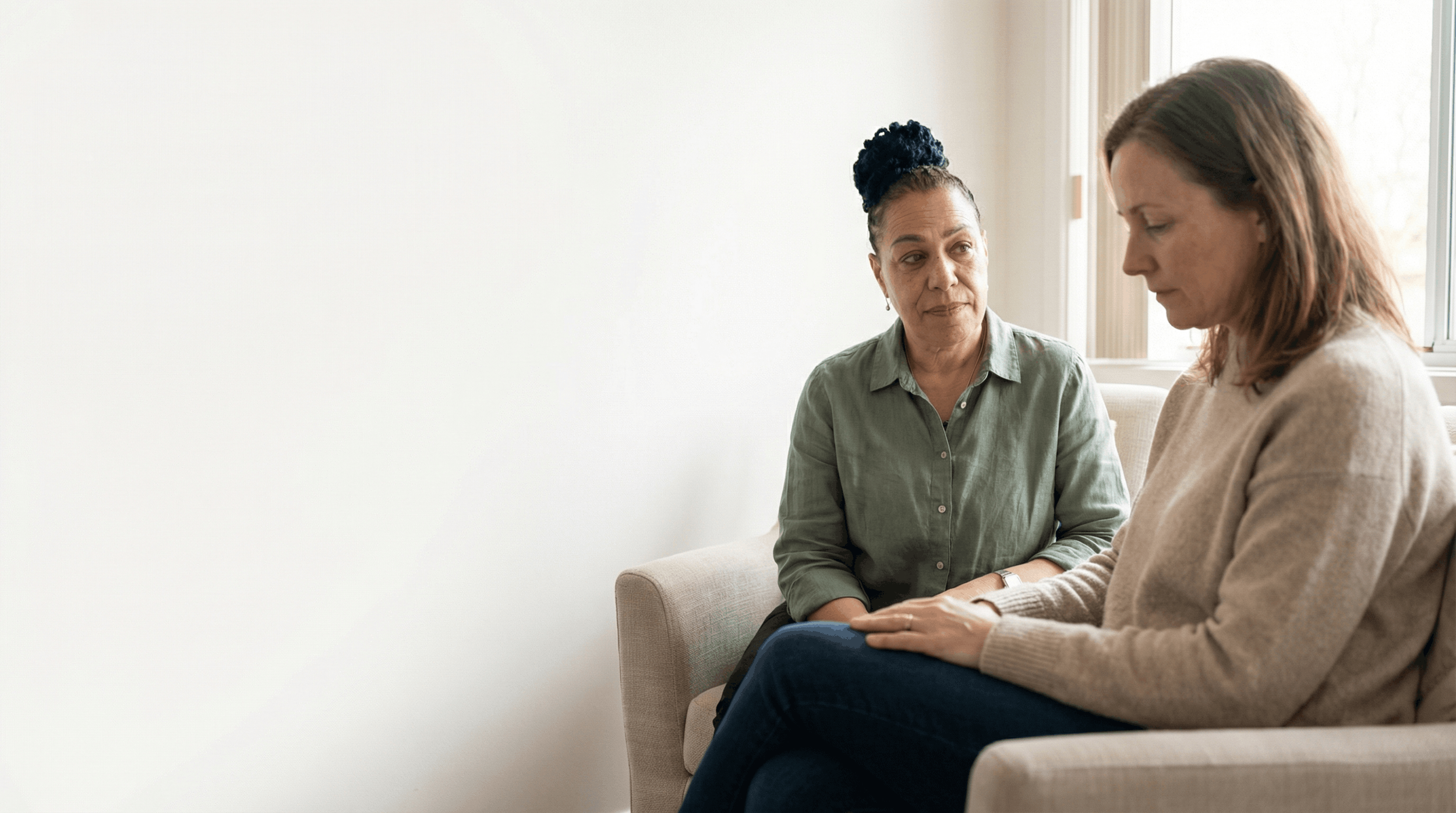 Two women engaged in a serious conversation sitting on a beige sofa in a bright room.