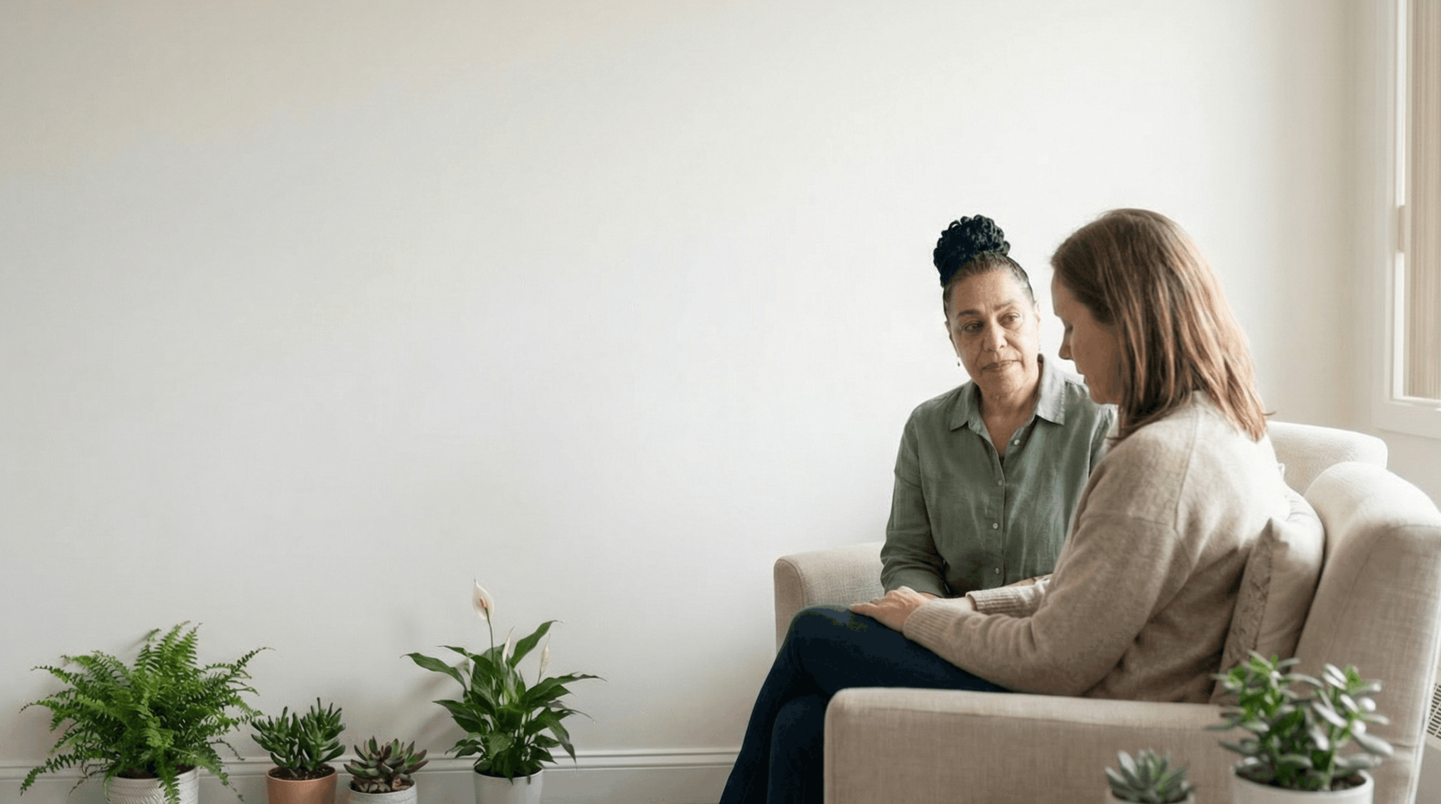 Two women sitting on a beige couch in a living room, having a conversation. There are several potted plants on the floor in front of them.