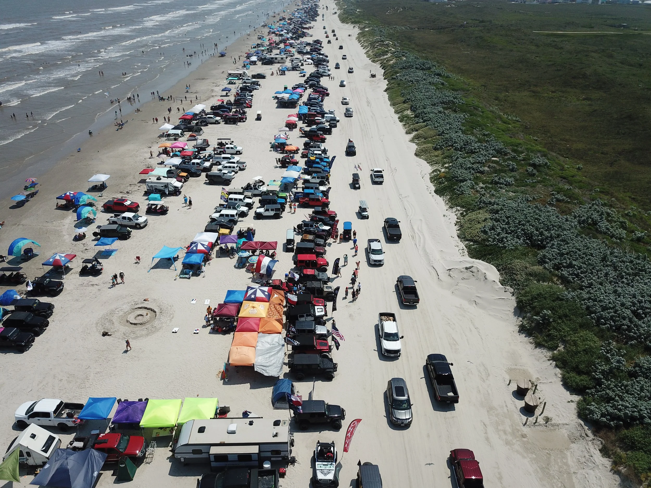 A crowded beach with numerous vehicles parked along the sandy shoreline and people enjoying the water and beach activities, with a green shrub-covered area on the right side.