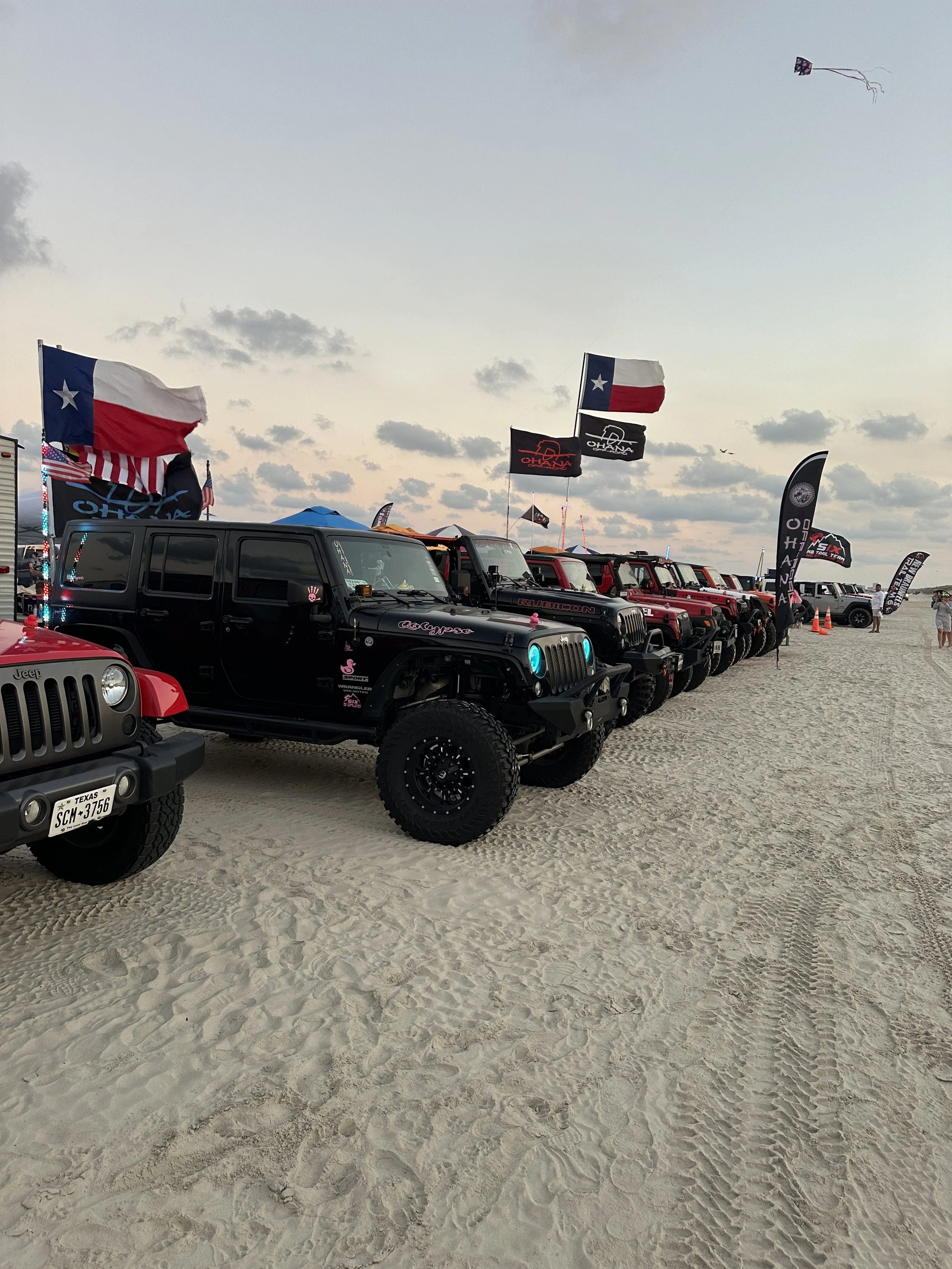 Several off-road vehicles, including a black Jeep, are parked on a sandy beach during a sunset or late afternoon, with Texas flags and festival banners flying overhead.