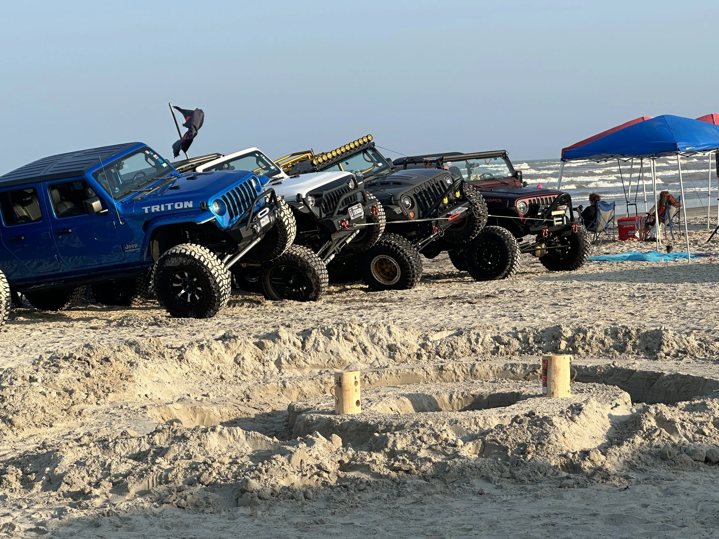 A row of modified off-road Jeeps parked on a sandy beach, with a canopy and people relaxing nearby by the ocean.