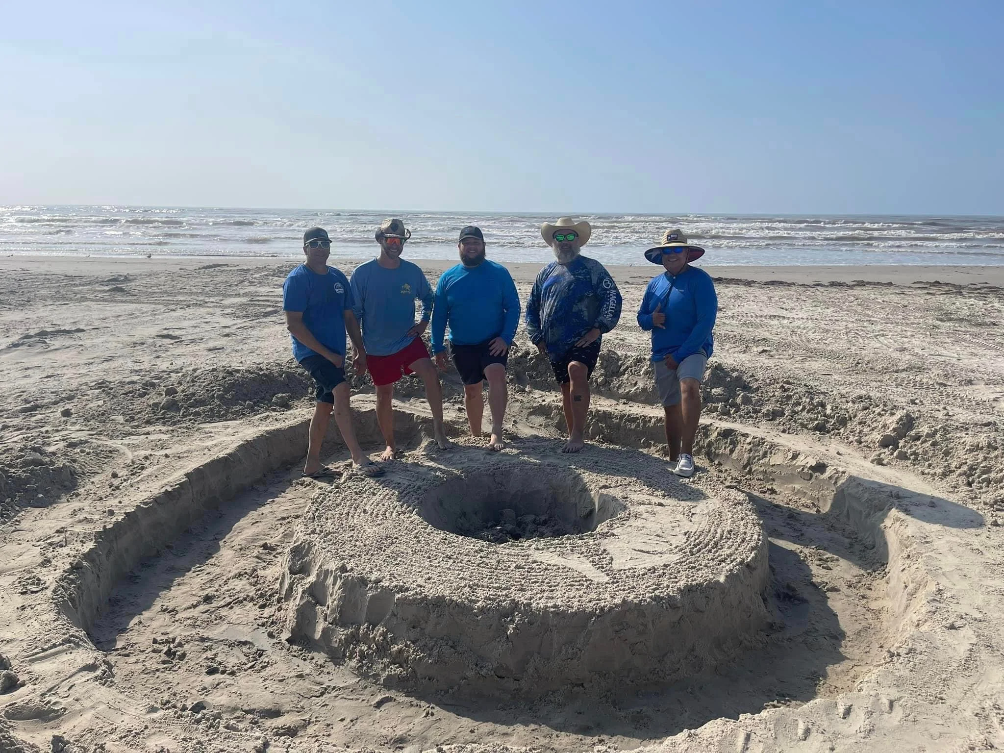 Five men standing on a beach with a sand sculpture of a large spiral or whirlpool design. The men are dressed in casual beach clothing with some wearing hats and sunglasses. The ocean and sky are visible in the background.