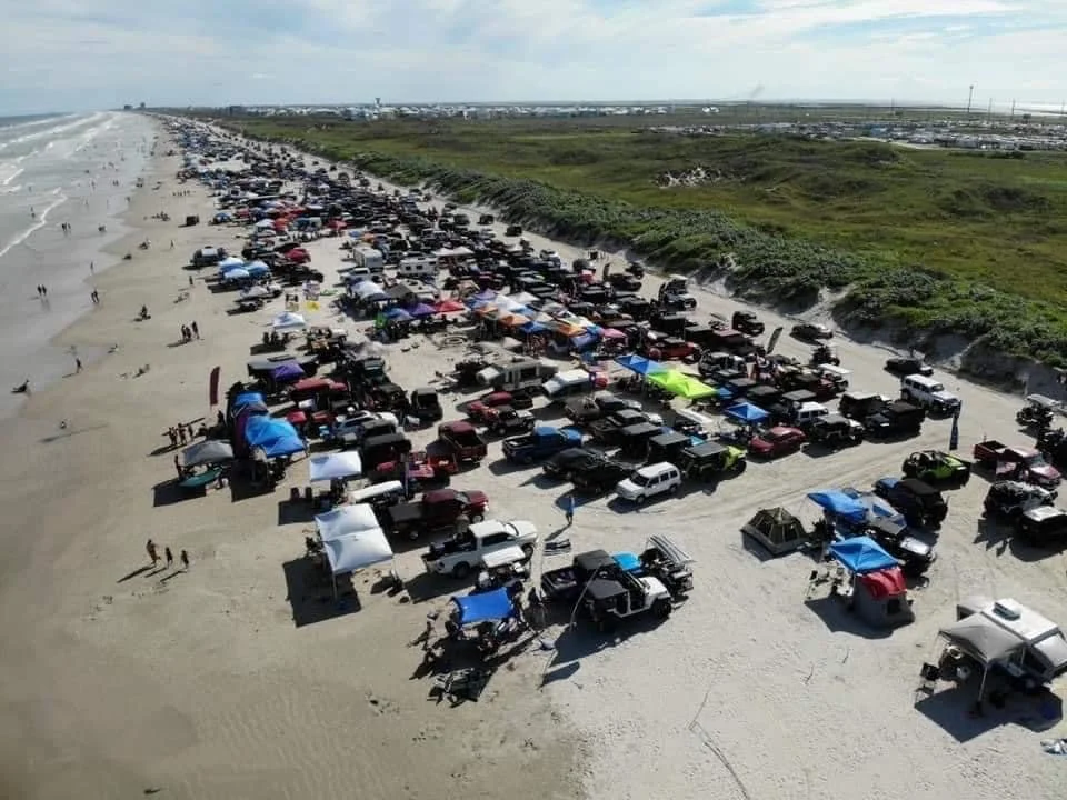 A crowded beach with numerous parked cars and tents along the shoreline, with people walking and enjoying the beach environment, green dunes in the background.
