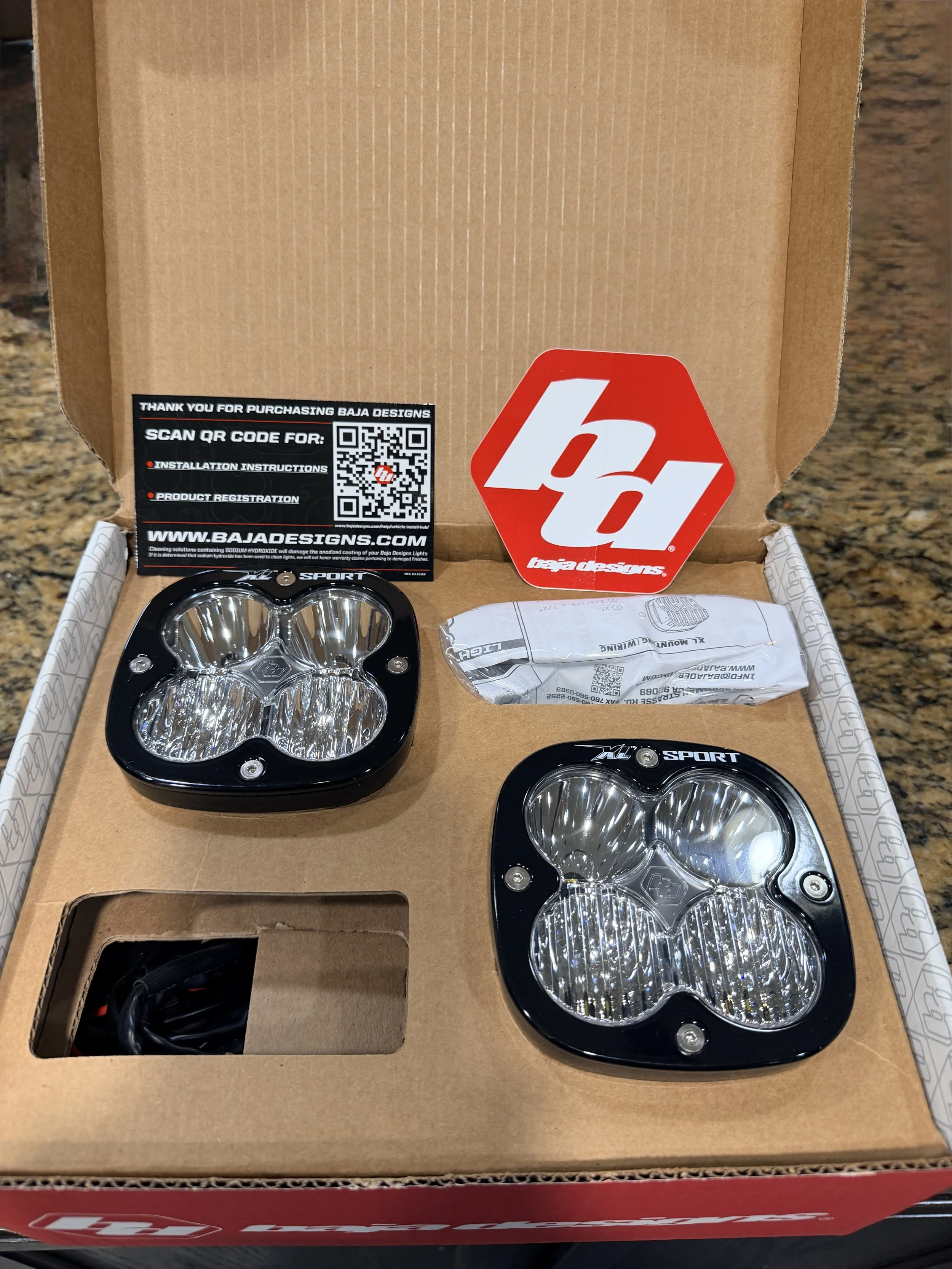 Box containing two black and clear LED headlights, a QR code sticker, and a Baja Designs logo, with a piece of plastic packaging and an instruction card inside a brown box.