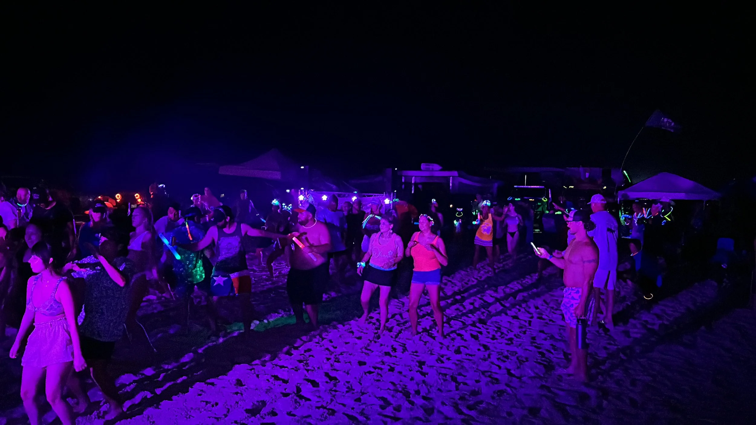Nighttime beach scene with a group of people dancing and socializing under blacklights and UV lights, with sand visible in the foreground.