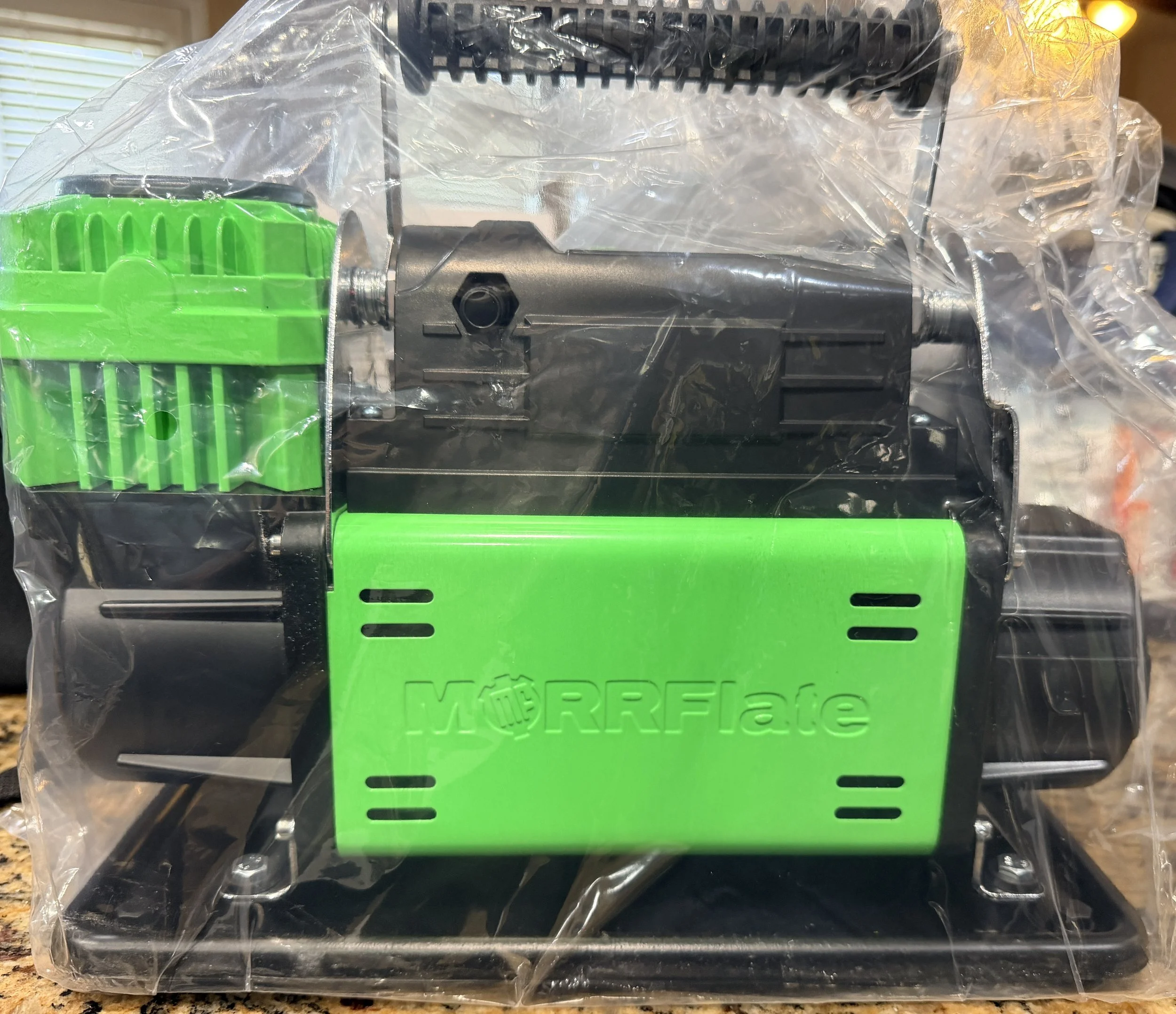 A power tool inside clear plastic wrapping, with a green and black body, on a countertop.