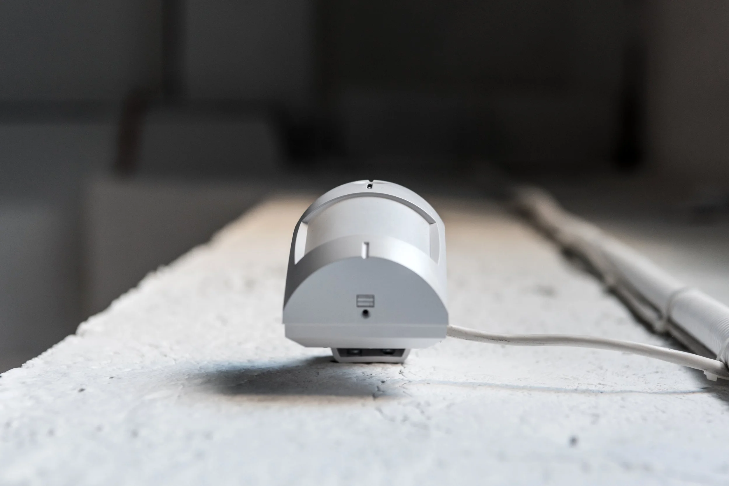 Close-up of a white electronic device with a power cord, placed on a white textured surface, with a blurred dark background.