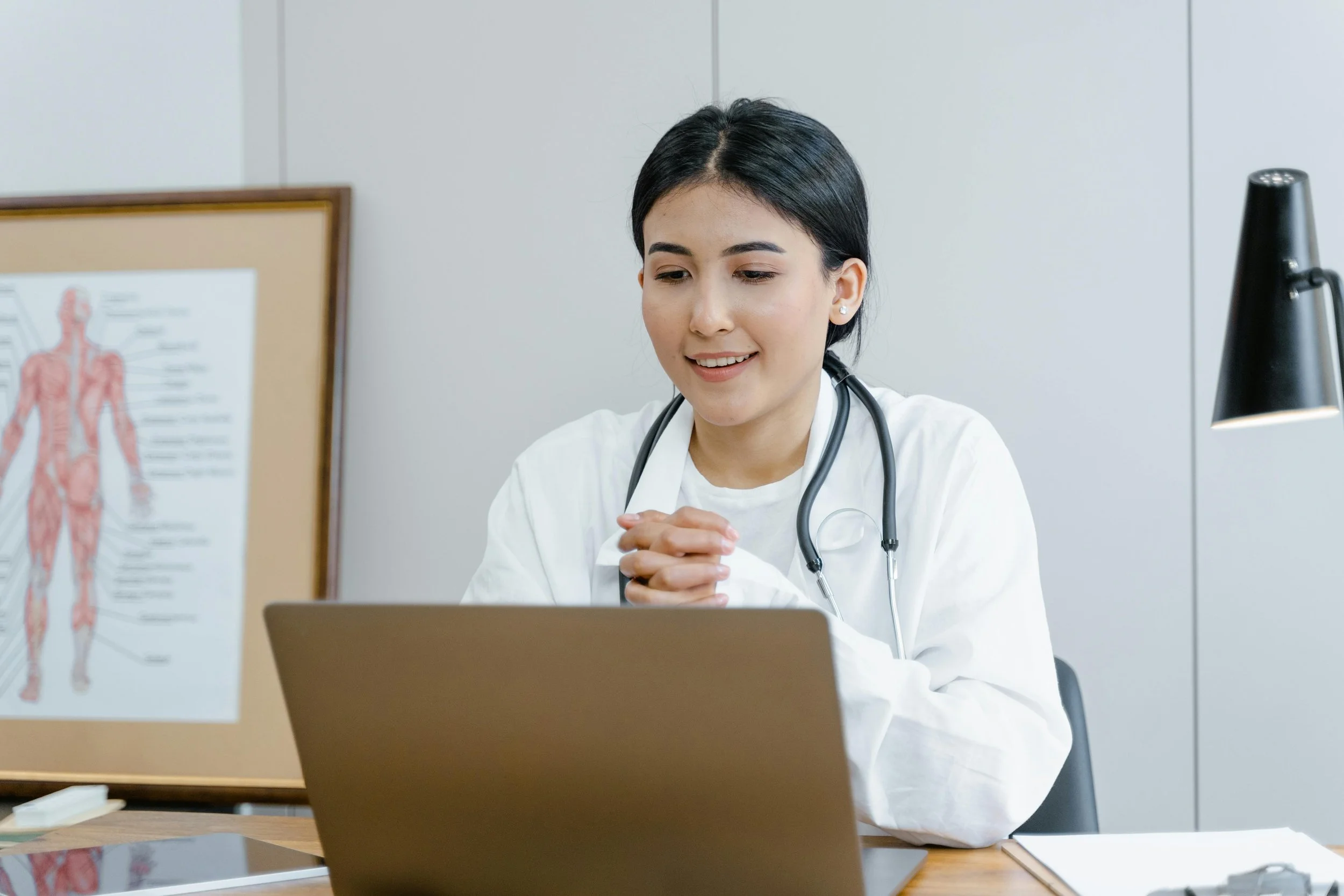 A female doctor with a stethoscope around her neck sitting at a desk and smiling during a video call, with a medical diagram of the human muscular system in the background.