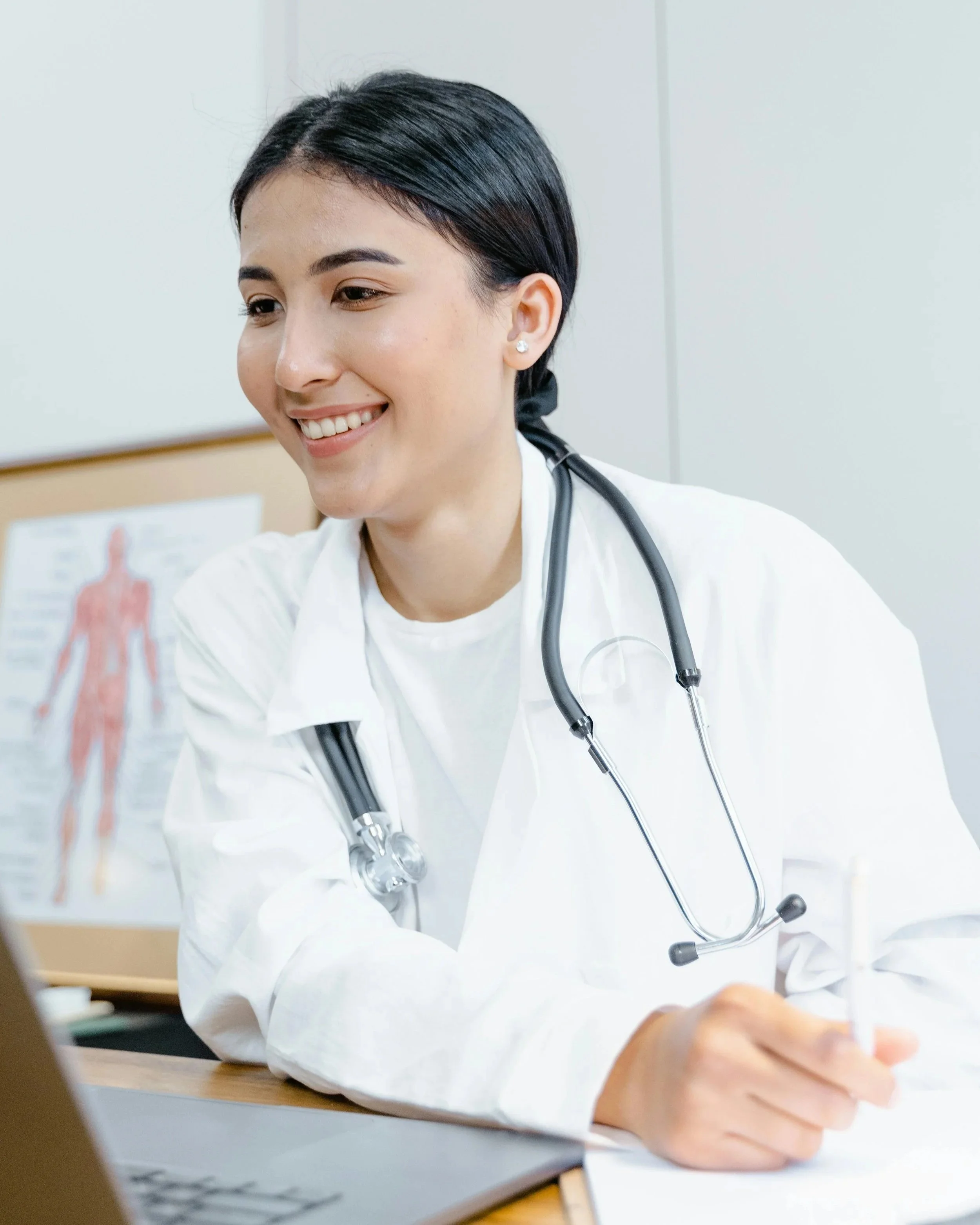 Smiling female healthcare professional with a stethoscope around her neck, writing on paper in an office or clinic setting.