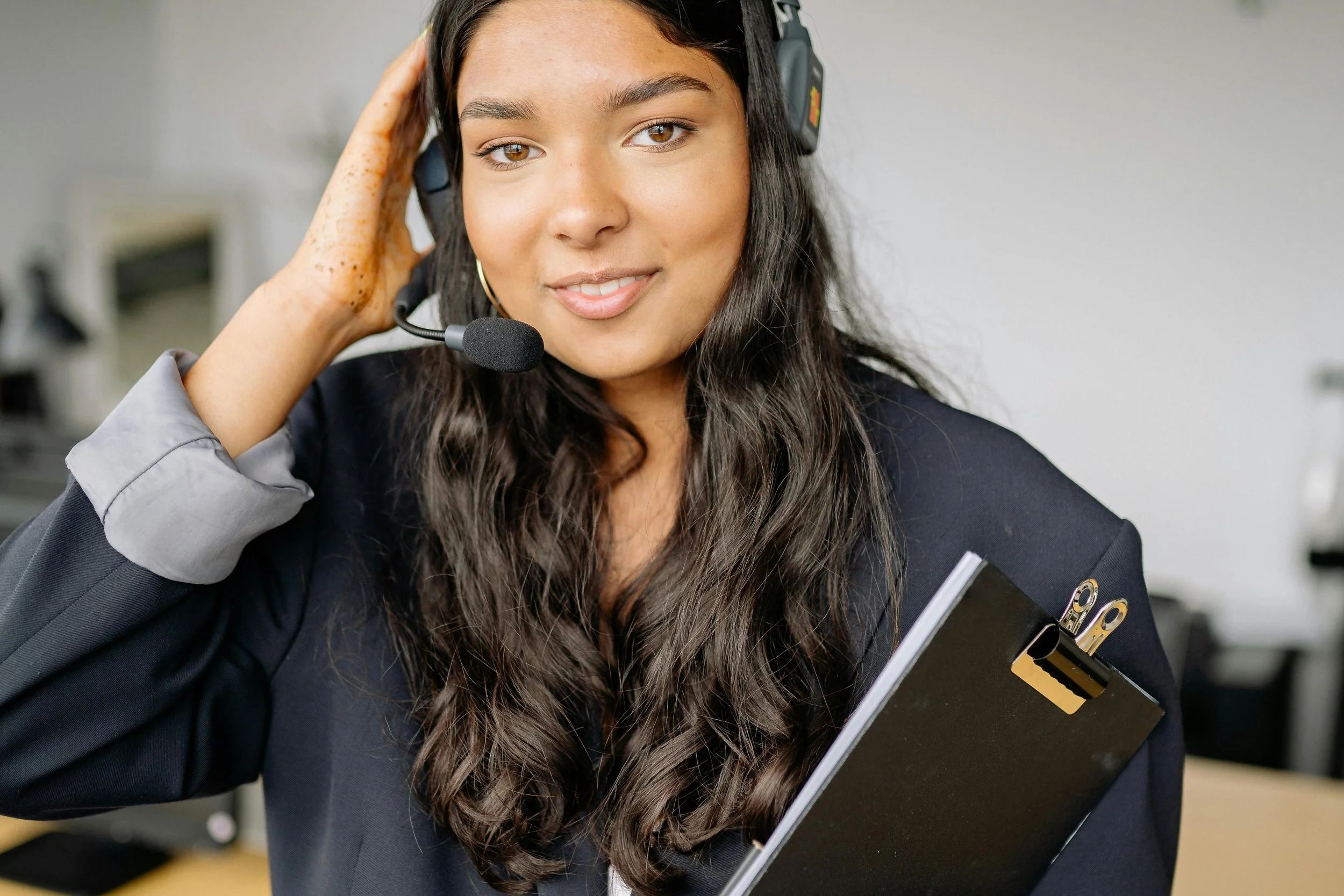 A young woman with dark hair wearing a headset and holding a clipboard, smiling at the camera.