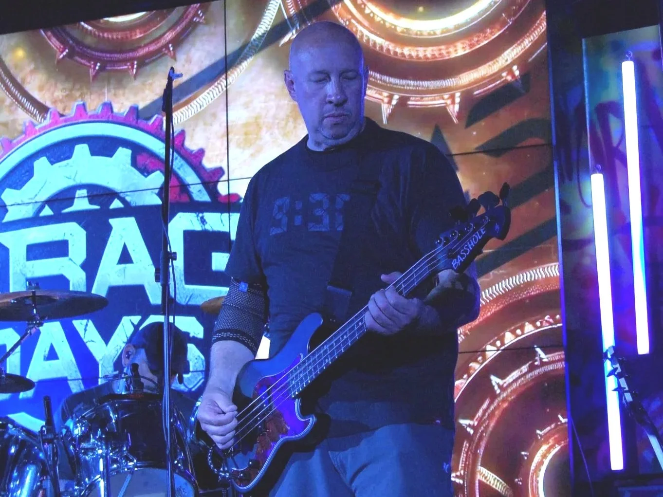 A man playing a bass guitar on stage with a colorful digital backdrop featuring large gears and the text "SPACE RAYS."