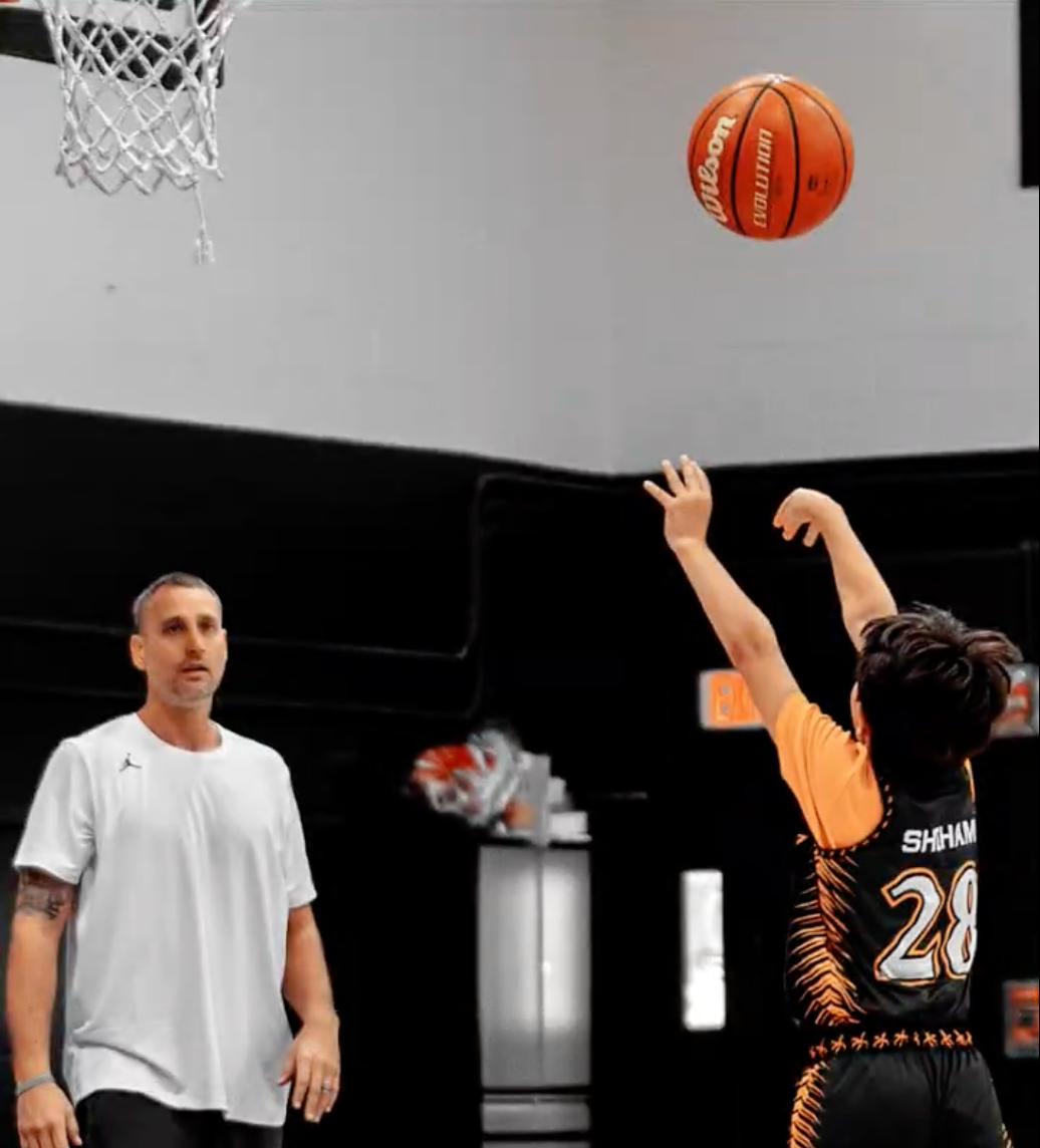A basketball shot in progress during a game or practice, with a player wearing a black and orange jersey numbered 28 shooting the ball towards the hoop, and a coach or referee watching nearby in a gym setting.