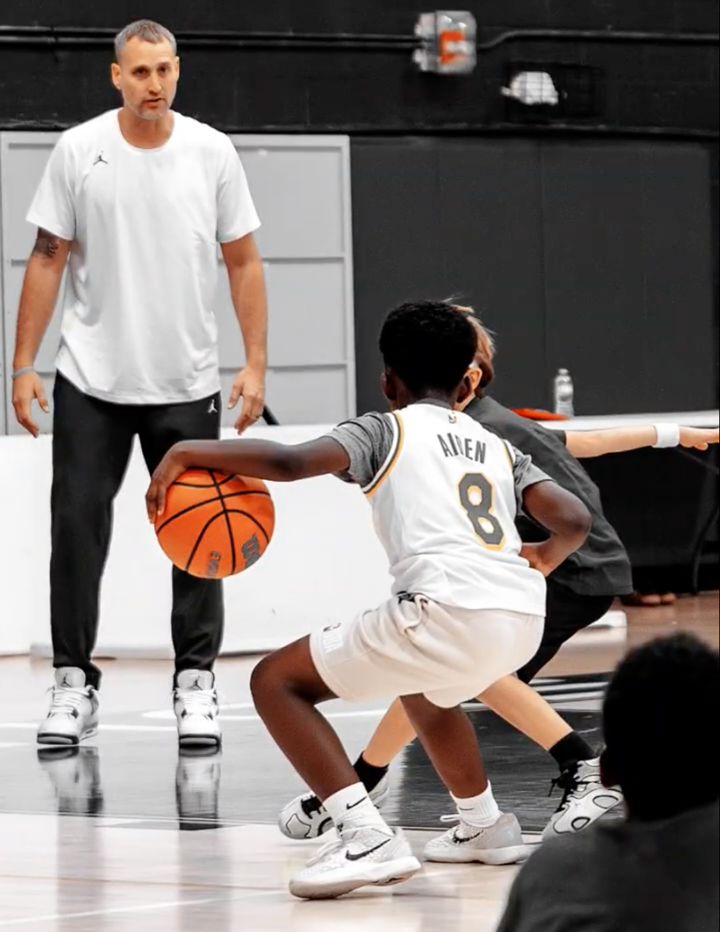 A young boy in a basketball uniform with the number 8, playing defense on a basketball court, with a person in black behind him and an adult coach watching in the background.