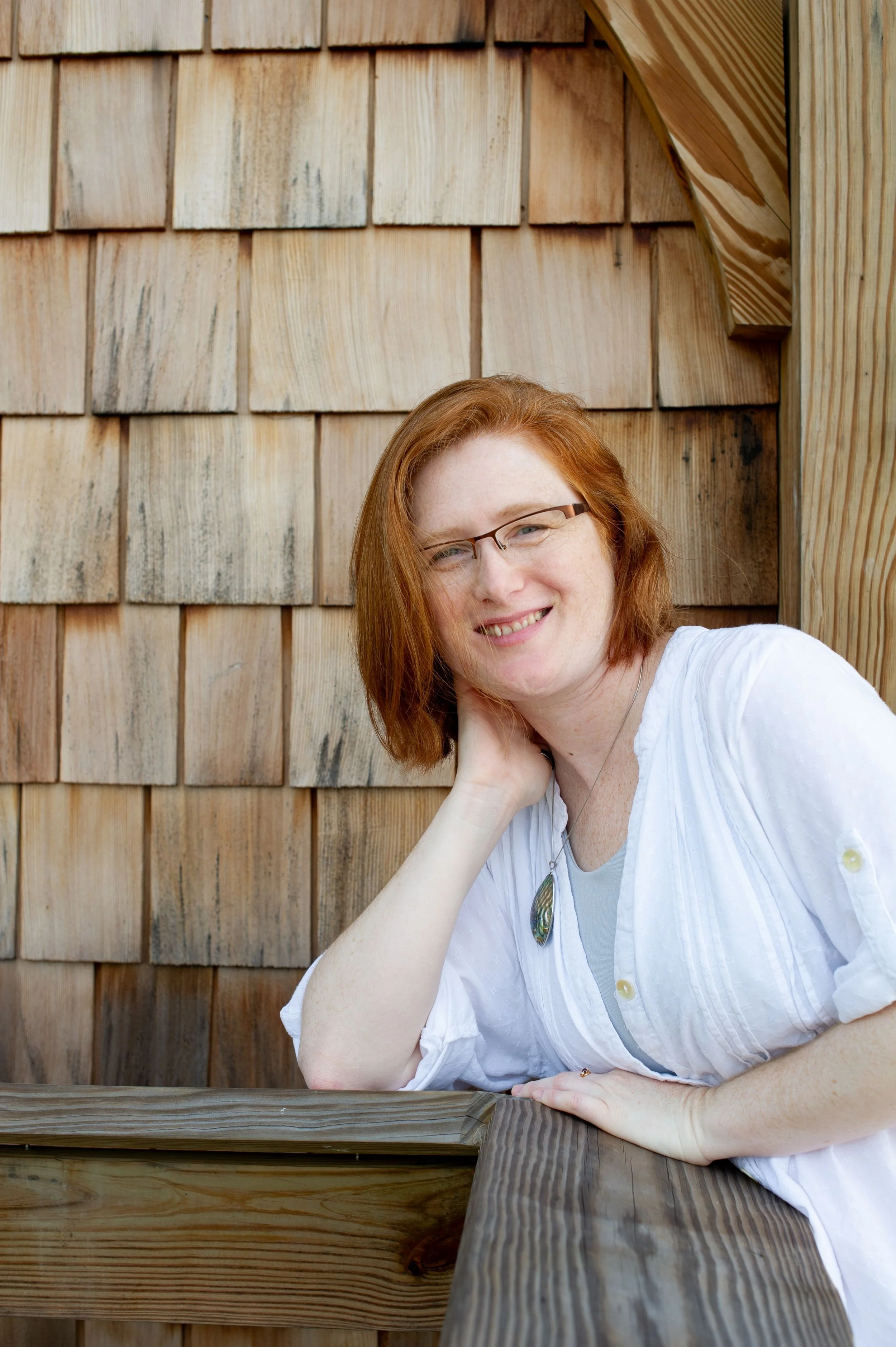 A woman with red hair and glasses leaning on her left hand, smiling at the camera, in front of a wooden house with shingle siding.