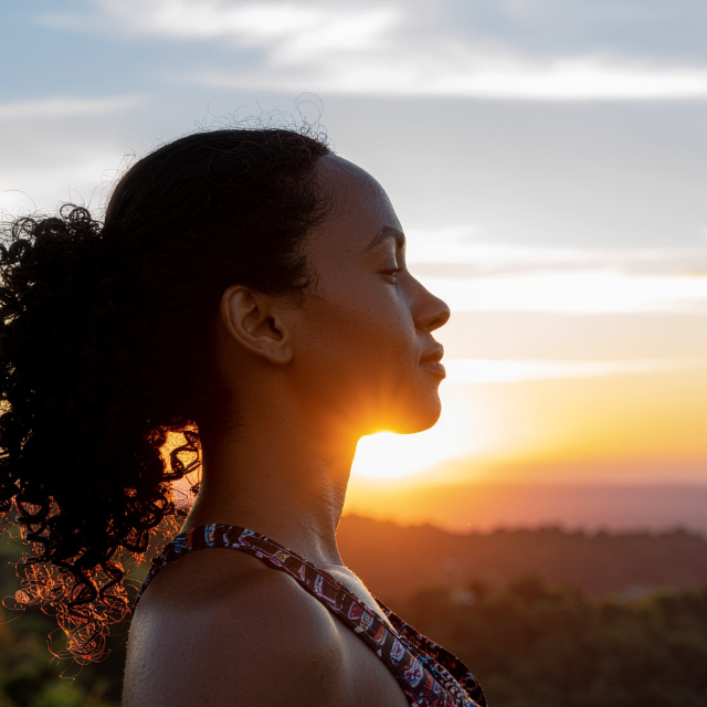 Side profile of a woman with curly hair in front of a sunset with a partly cloudy sky.