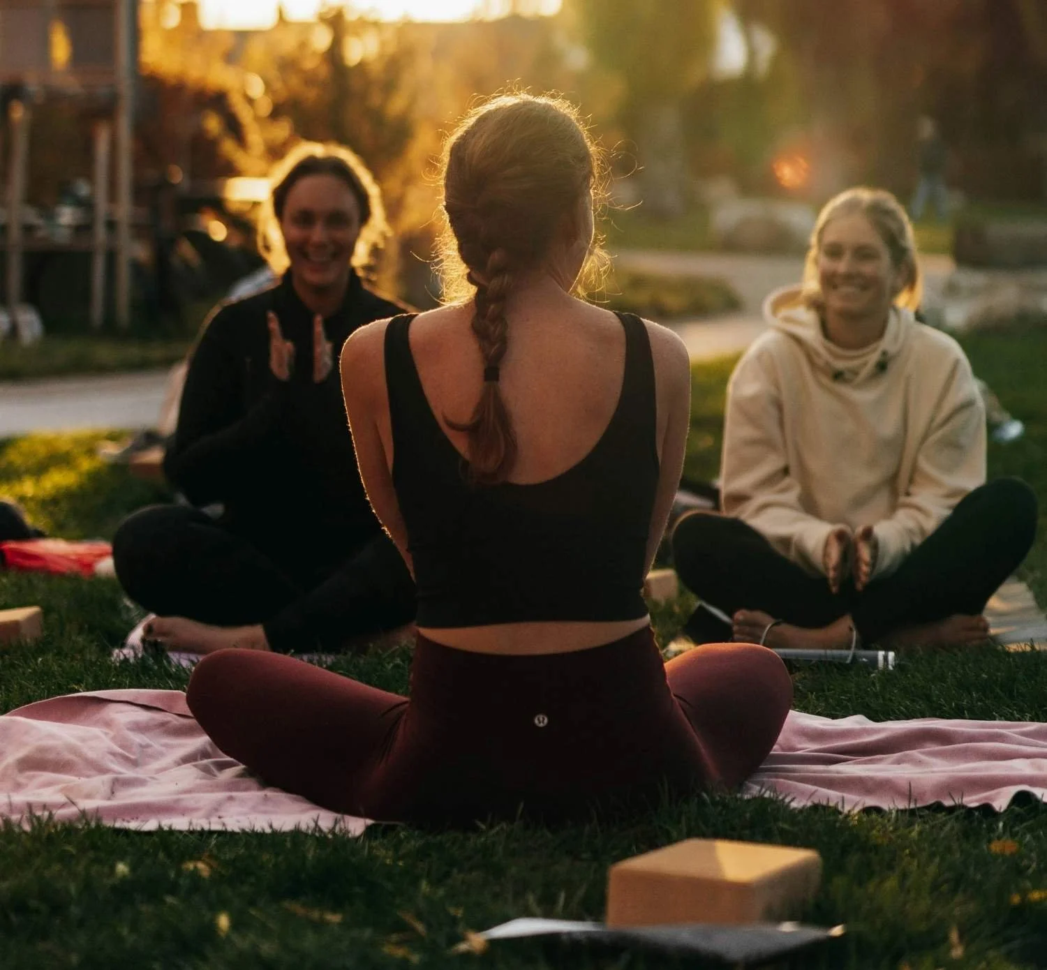 A group of women sitting on the grass in a park during sunset, practicing wellness & yoga.
