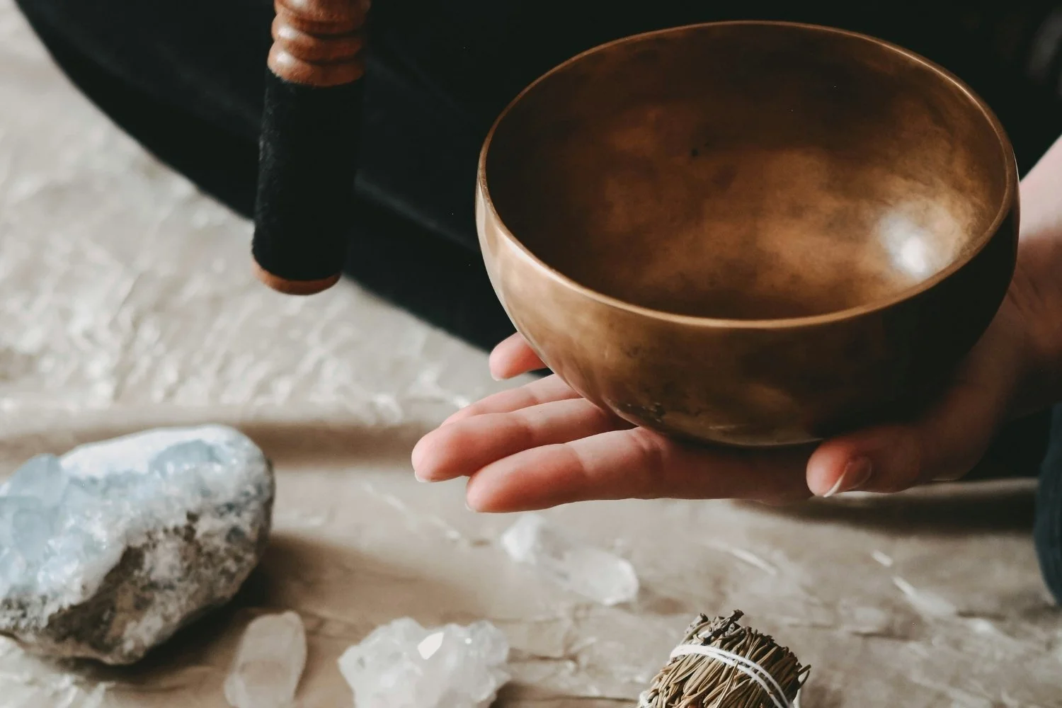 A person holding a metallic singing bowl with a dark interior, with various crystals and stones on a textured surface below.