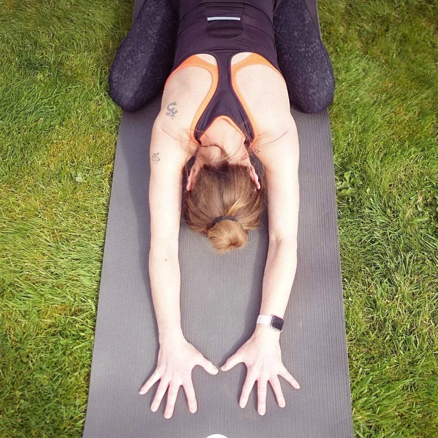 A woman lying face down on a yoga mat outdoors, stretching her arms overhead, on green grass.