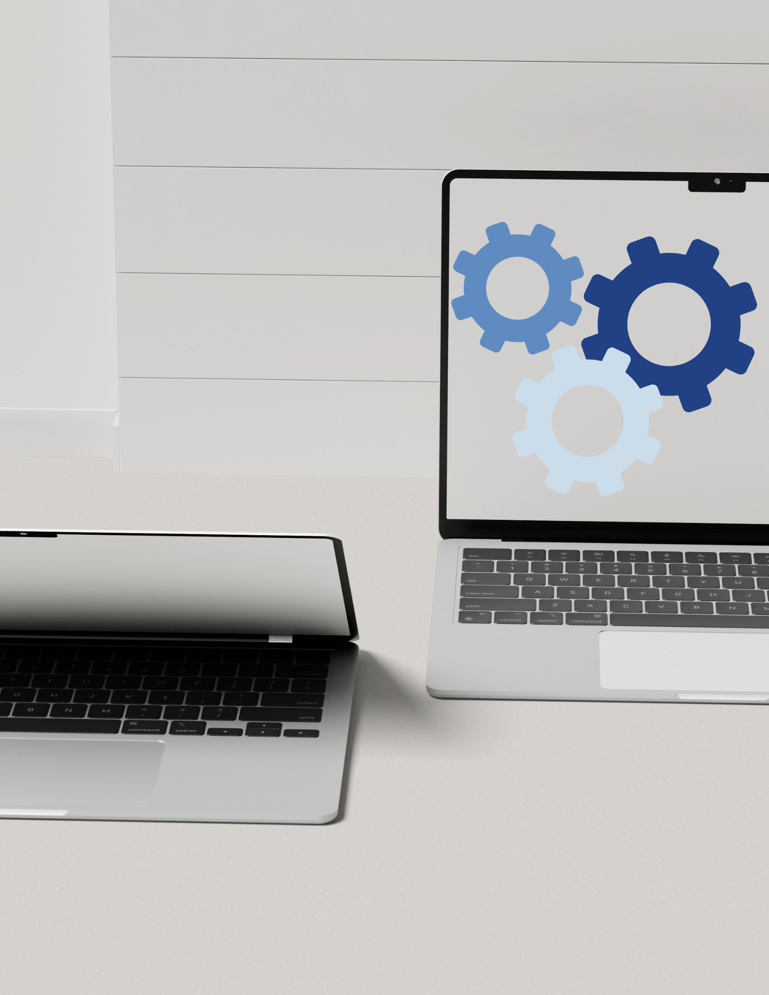 Two laptops on a white desk with a white wall in the background. The right laptop screen displays three gear icons in different shades of blue.