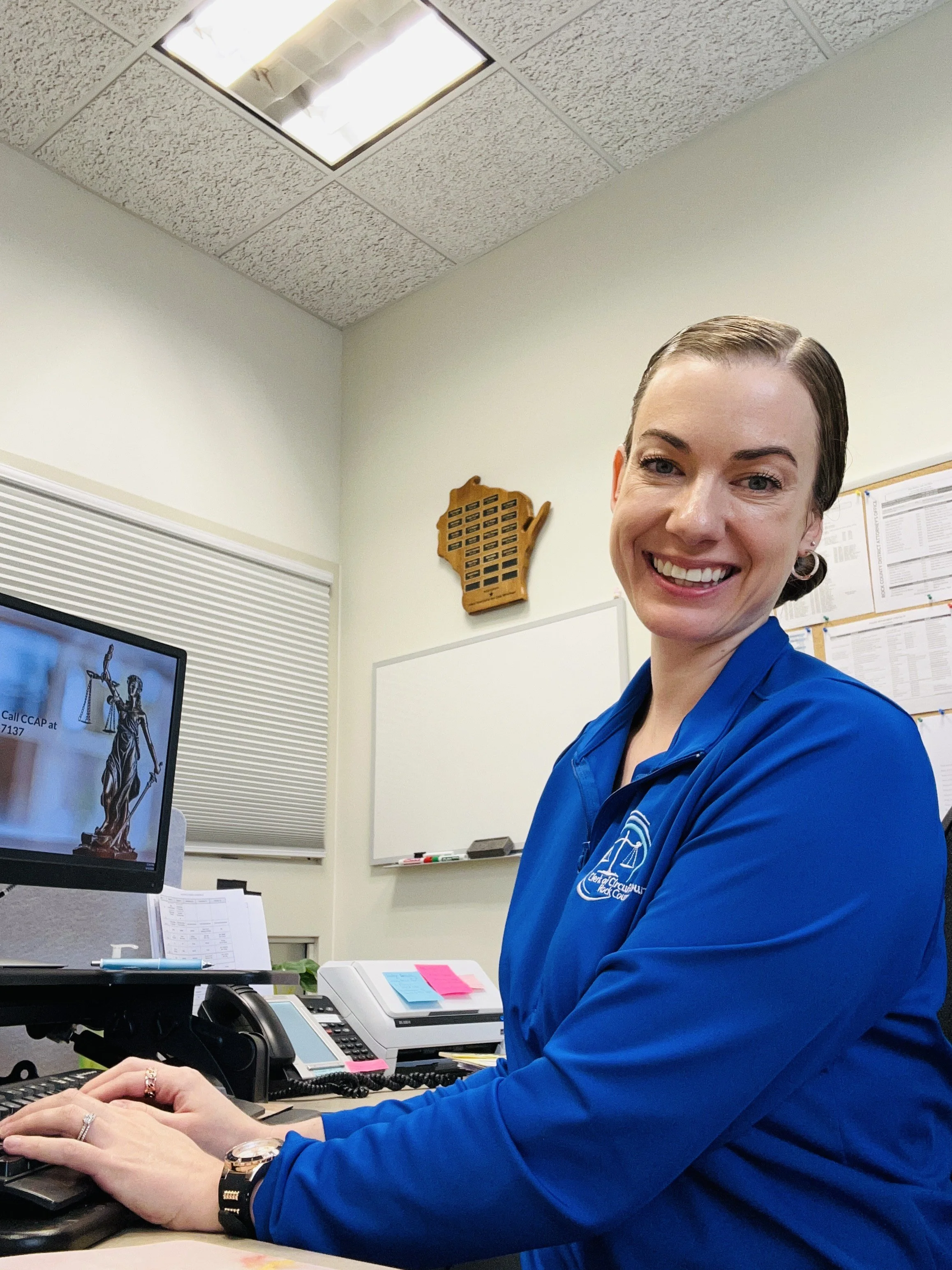 Amanda smiling in a blue jacket with the Clerk of Circuit Court logo sits at a desk in her office, working on a computer. There is a whiteboard, a decorative Wisconsin-shaped wood plaque, and a computer monitor on her desk.
