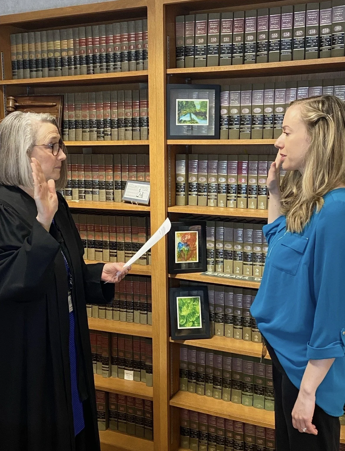Amanda taking the oath of office as Clerk of Circuit Court with her right hand raised while Honorable Barbara McCrory holds the oath document in front of her in a room with wood bookshelves filled with law books and framed artwork.