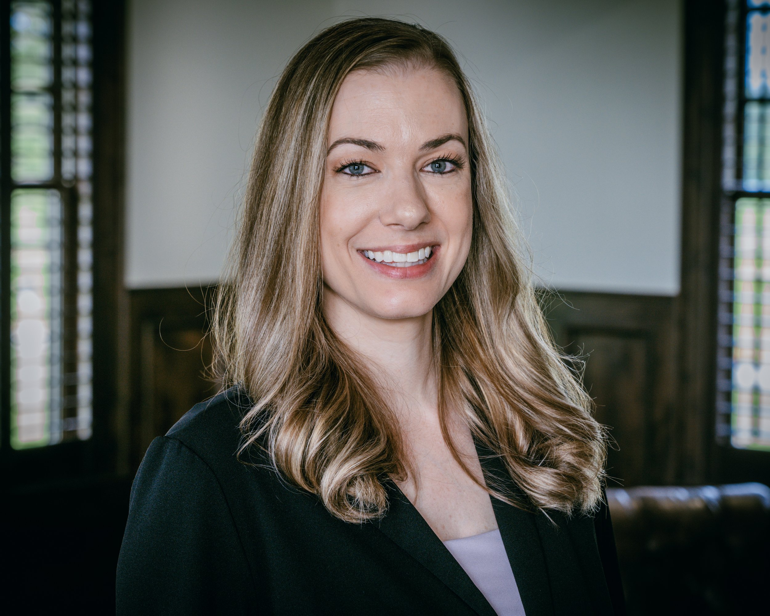 A headshop of Amanda. She has long, wavy blonde hair smiling in a room with wooden paneling and window shutters.