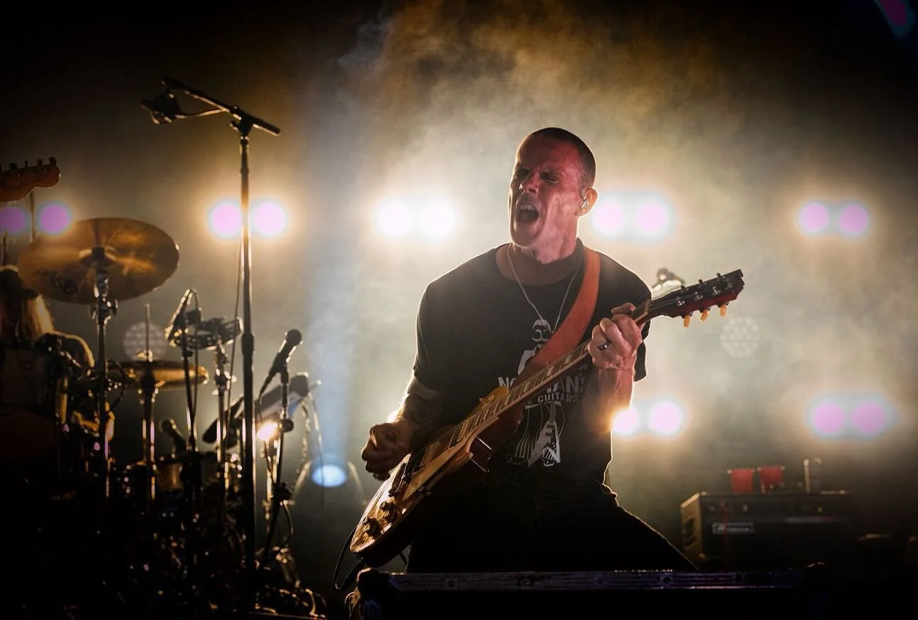 A male guitarist passionately performs on stage, with bright backlighting and fog, accompanied by a drummer and musical equipment.