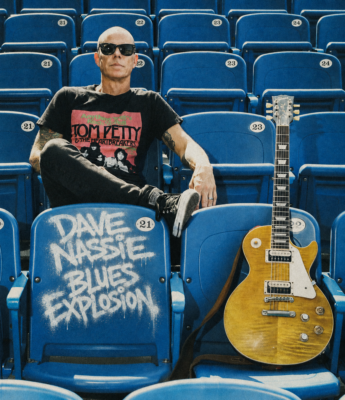 A bald man wearing sunglasses, a Tom Petty t-shirt, and black pants sitting in an empty blue stadium seat. There is an electric guitar resting on a nearby seat with graffiti that reads "DAVE NASSIE BLUES EXPLOSION."
