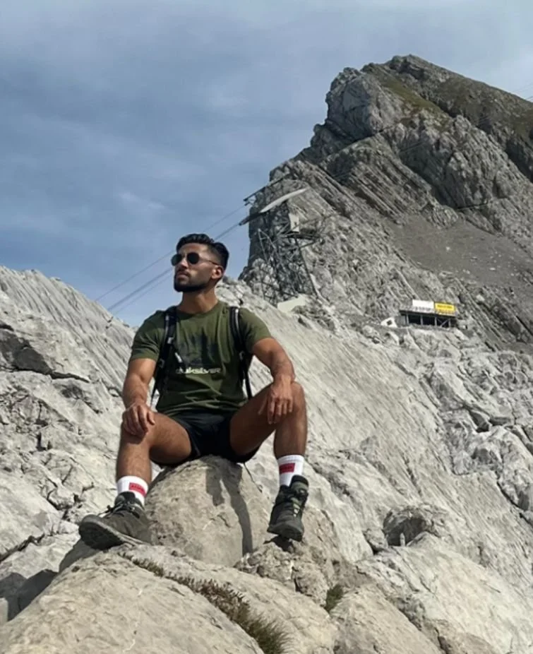 Ein Mann sitzt auf großen Steinen in einer felsigen Bergregion, trägt eine grüne T-Shirt, schwarze Shorts, Wanderschuhe, Sonnenbrille und einen Rucksack, im Hintergrund ist eine Seilbahn und ein Bauwerk sichtbar.