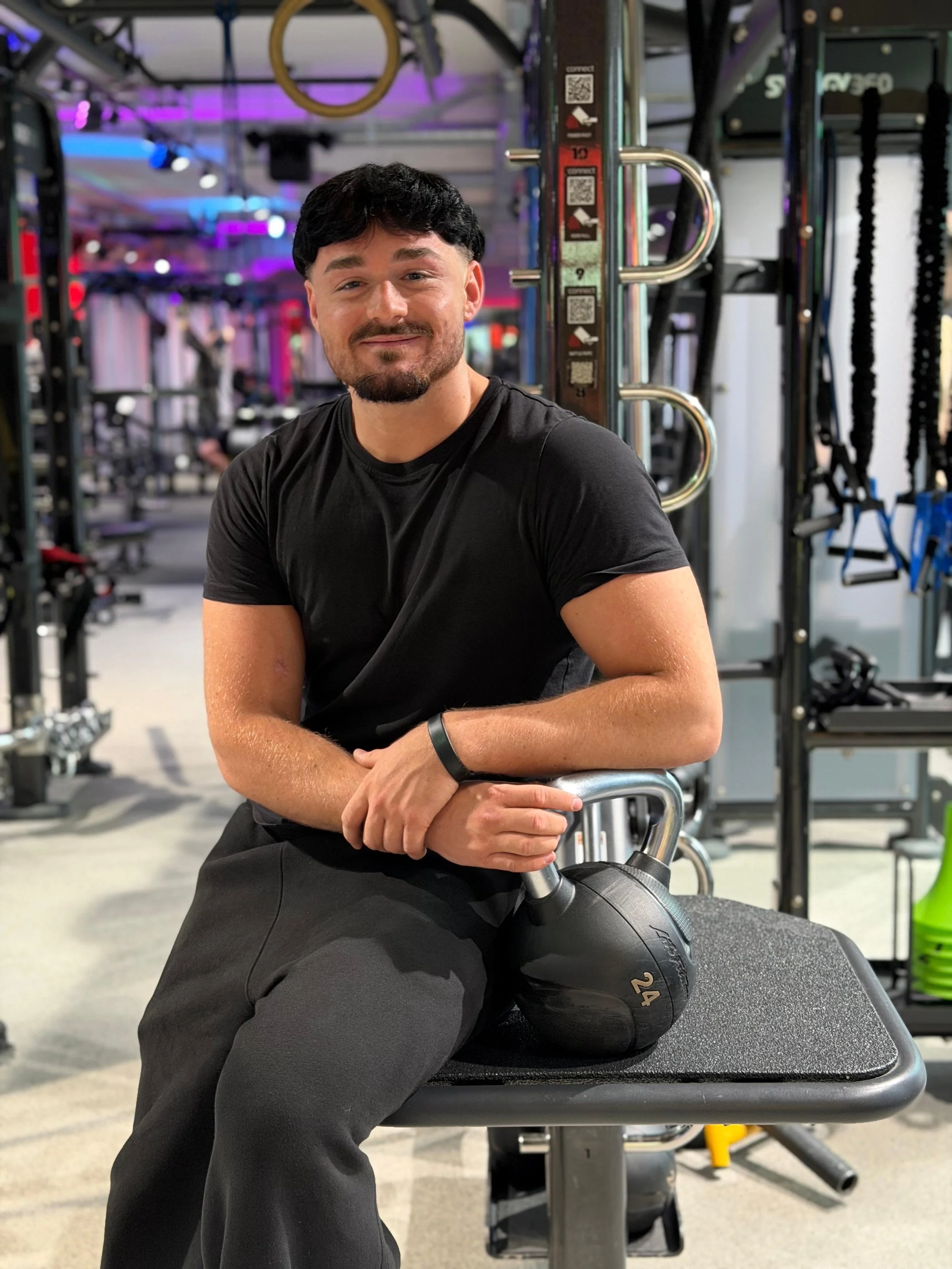 Ein Mann sitzt in einem Fitnessstudio, hält eine Kettlebell mit der Hand, trägt ein schwarzes T-Shirt und schwarze Hosen, im Hintergrund sind Fitnessgeräte und bunte LED-Lichter.