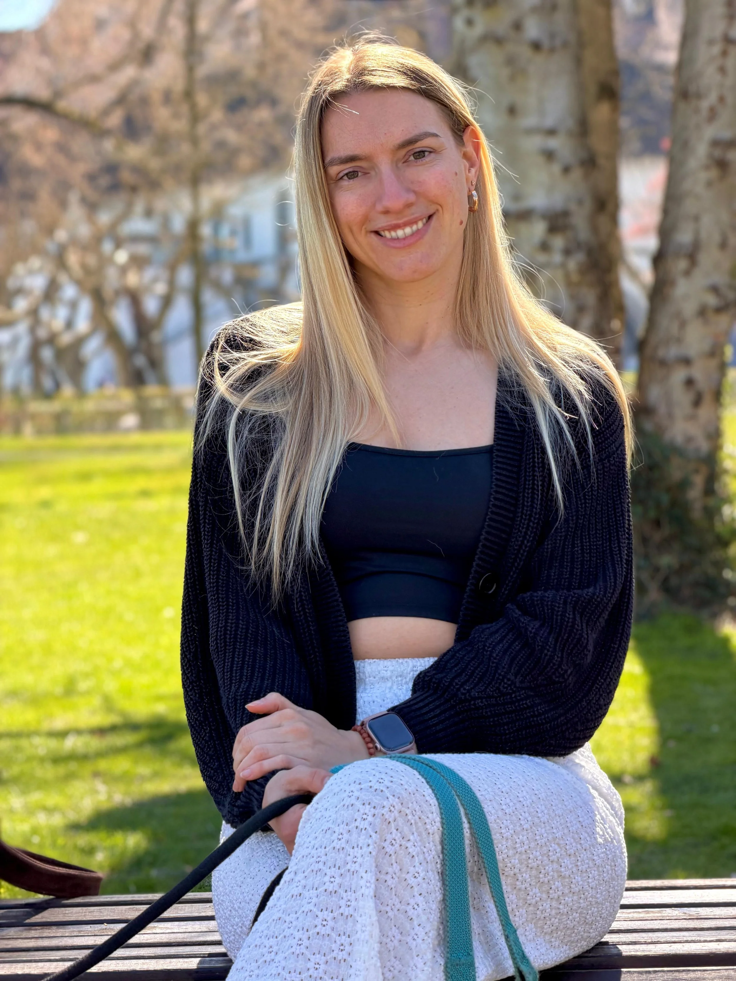 Junge Frau mit langen blondem Haar, Sonnenbrille, schwarzem Crop-Top, schwarzer Strickjacke und weißen Hosen sitzt auf einer Parkbank im Sonnenschein, umgeben von Bäumen.