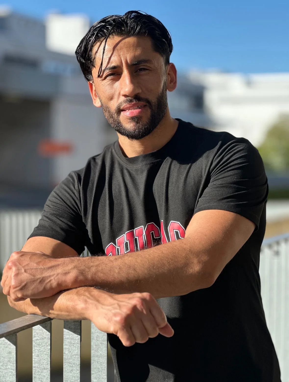 Mann mit dunklen Haaren und Bart trägt schwarzen T-Shirt mit roter Aufschrift, steht im Freien bei Sonnenschein, lehnt an einem Geländer, im Hintergrund ein Gebäude und blauer Himmel.