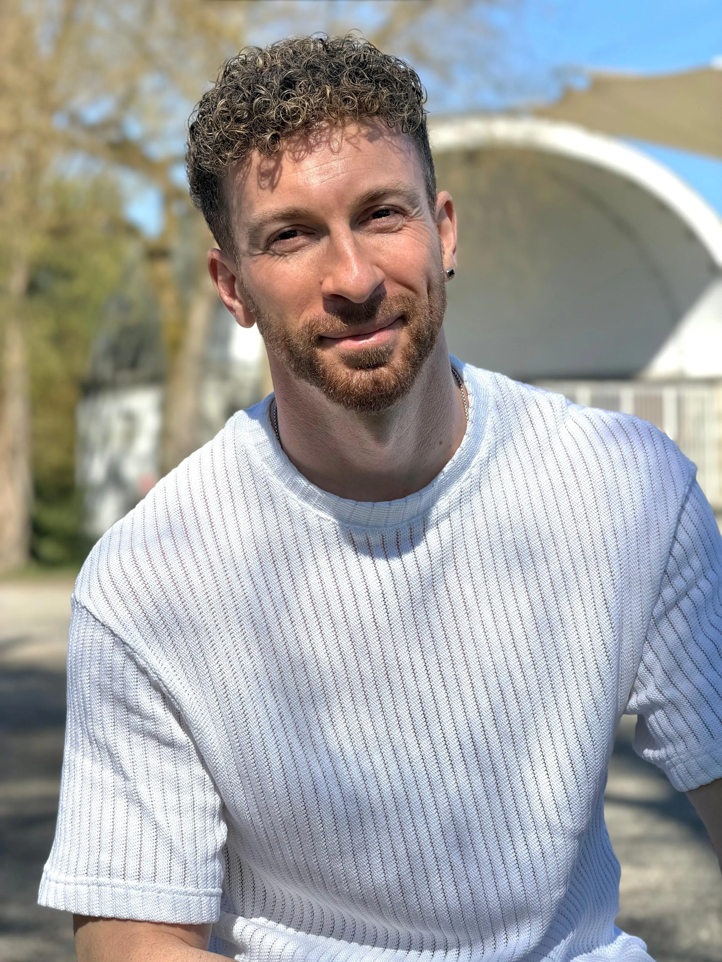 Ein junger Mann mit kurzen, lockigen Haaren und einem kurzen Bart, trägt ein weißes, gestreiftes T-Shirt, steht im Freien bei sonnigem Wetter, im Hintergrund sind Bäume und eine weiße, gebogene Struktur sichtbar.