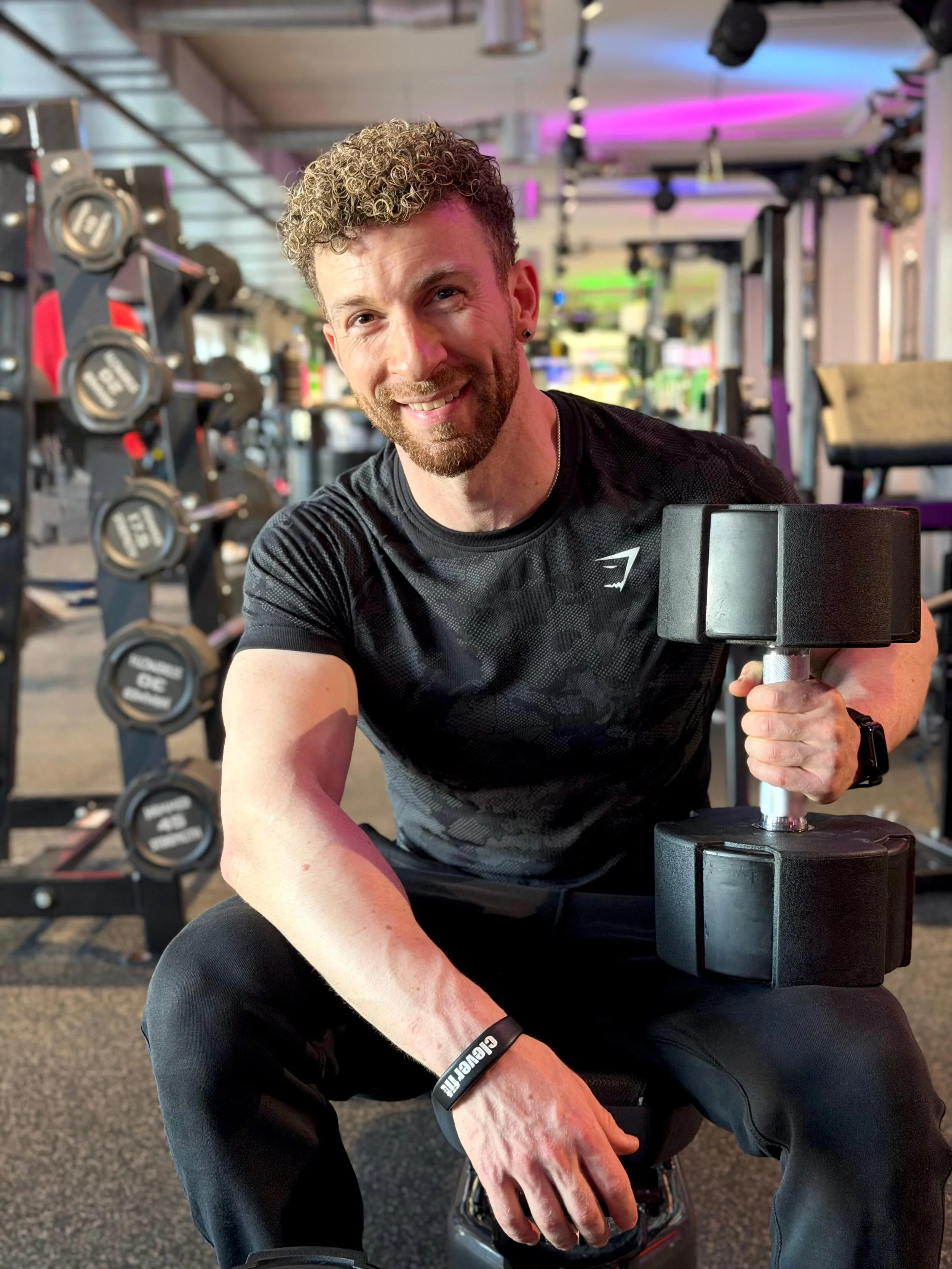 Mann im Fitnessstudio, der mit einem schwarzen Gummiband trainiert, in der Hand ein schwarzes Gewicht haltend und lächelnd in die Kamera schauend.