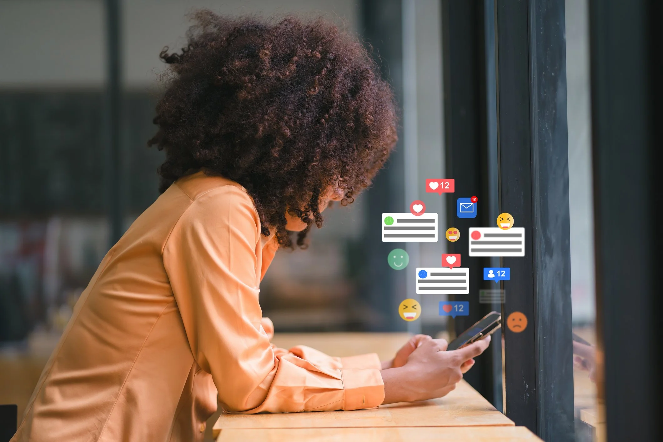 A woman with curly hair in a peach-colored shirt is using a smartphone near a window. Floating social media icons, including likes, messages, and reactions, surround her.