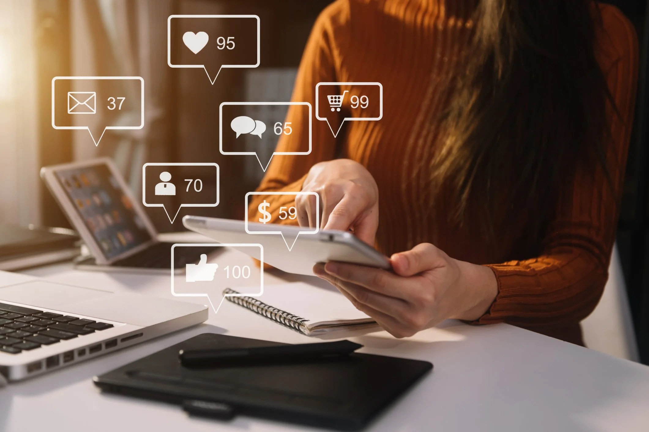 Person using a smartphone surrounded by floating icons representing social media interactions, messages, likes, shopping, and comments, in a workspace with a laptop, notebook, and tablet.