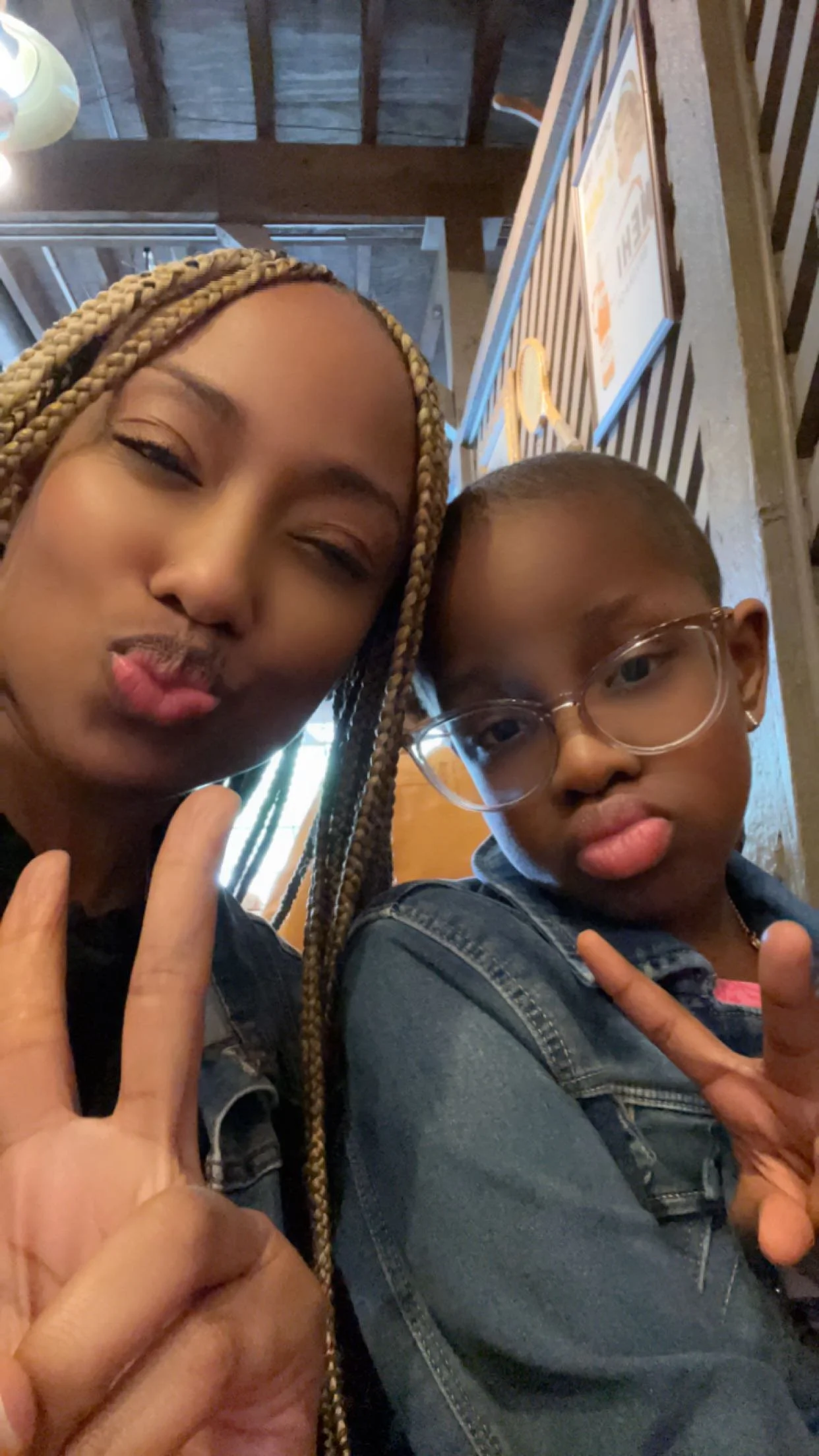 Shelby and her mom taking a selfie together inside a restaurant or cafe, both making peace signs with their hands. The woman has long blonde braids and the girl has short hair, glasses, and is wearing a denim jacket.