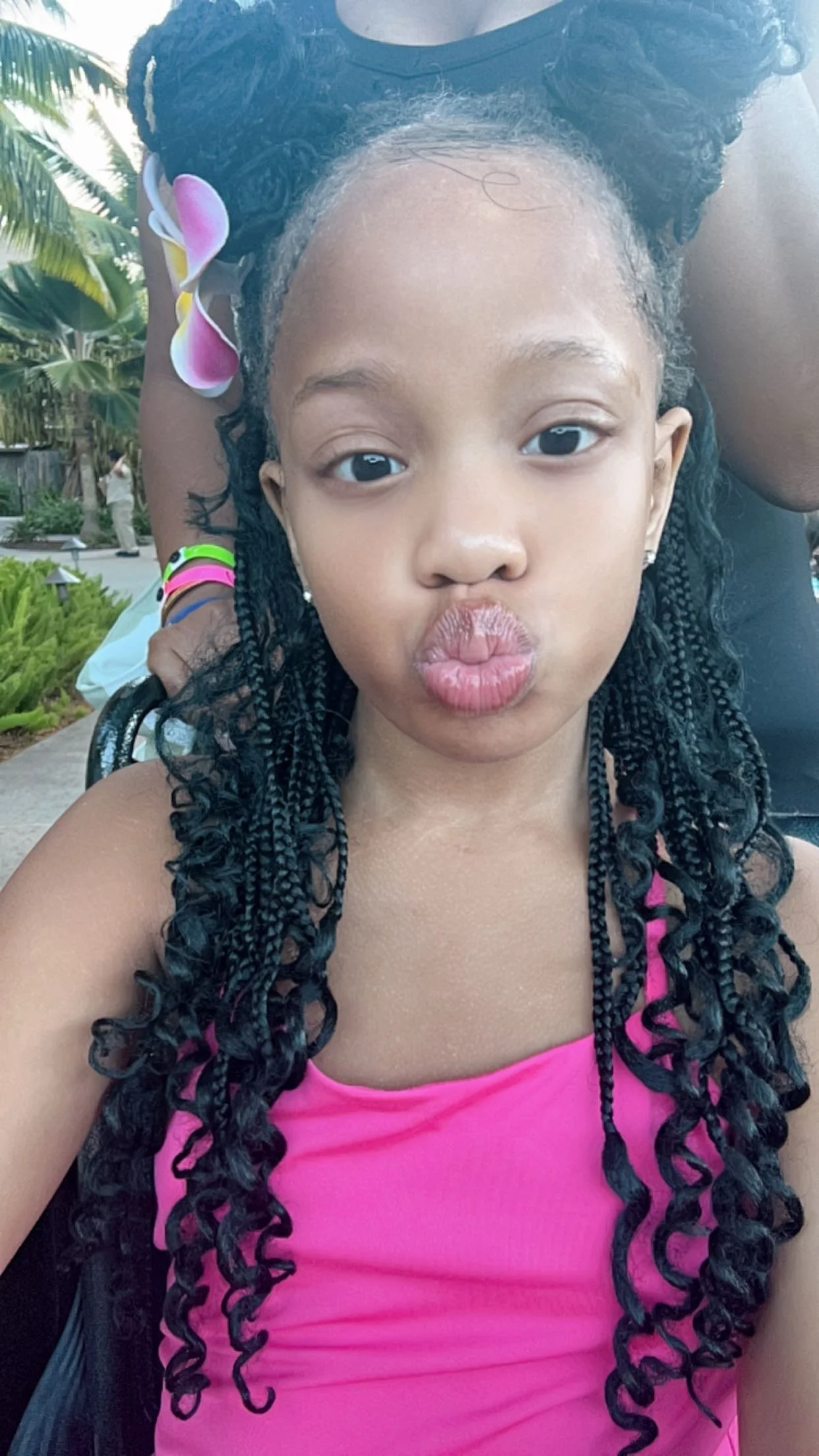 Young girl with long, curly black hair making a duck face for the camera, wearing a pink top, with a flower clip in her hair and earrings, outdoor setting with palm trees and greenery in the background.
